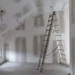 Wnętrze w trakcie prac wykończeniowych: ściany i sufit pokryte płytami gipsowo-kartonowymi, widoczne łączenia szpachlowane, aluminiowa drabina rozstawiona w centrum pomieszczenia, małe okno...