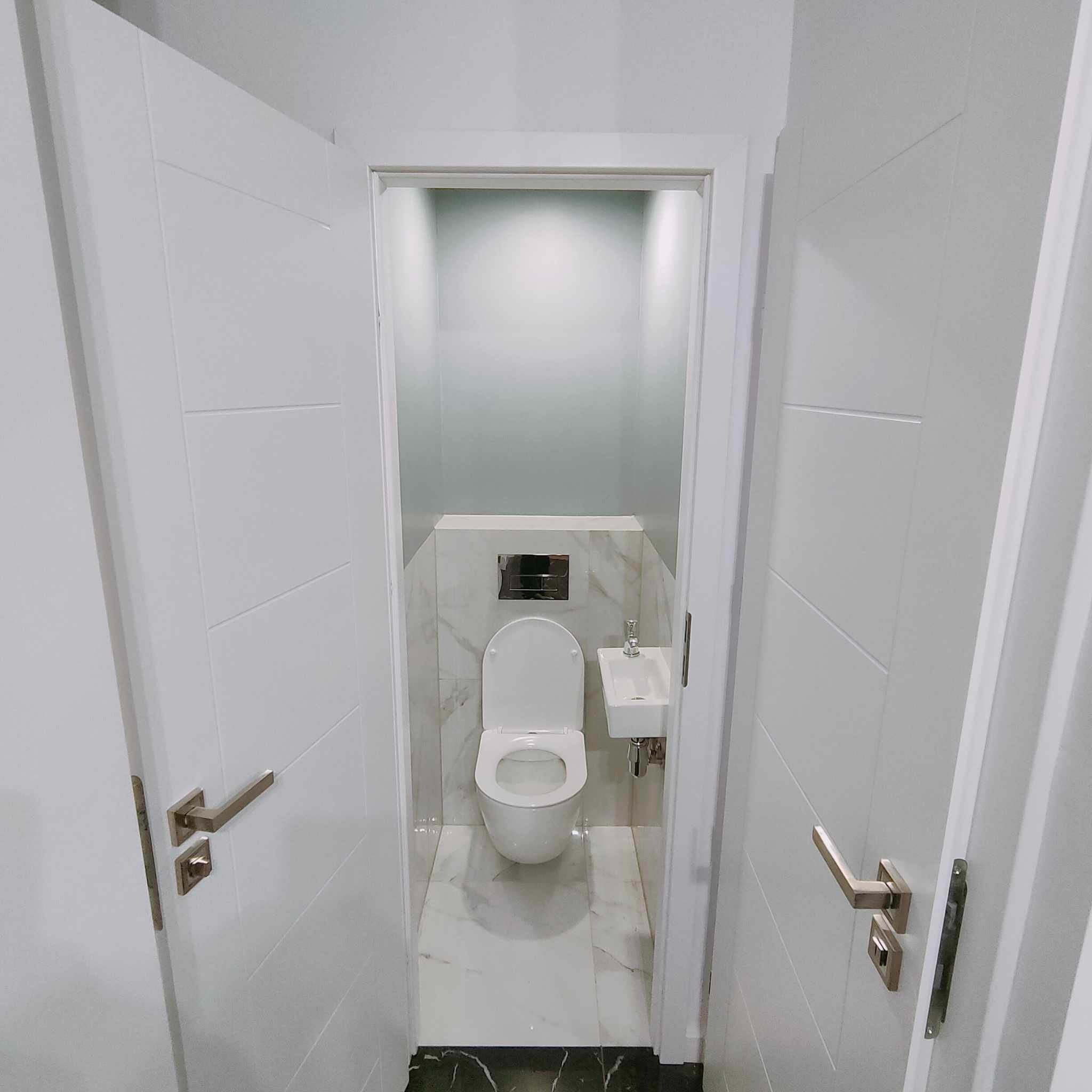 Mała, nowoczesna toaleta z podwieszaną miską WC, umywalką nablatową i białymi marmurowymi płytkami na ścianach, widziana z perspektywy otwartych, białych drzwi z poziomymi frezami.