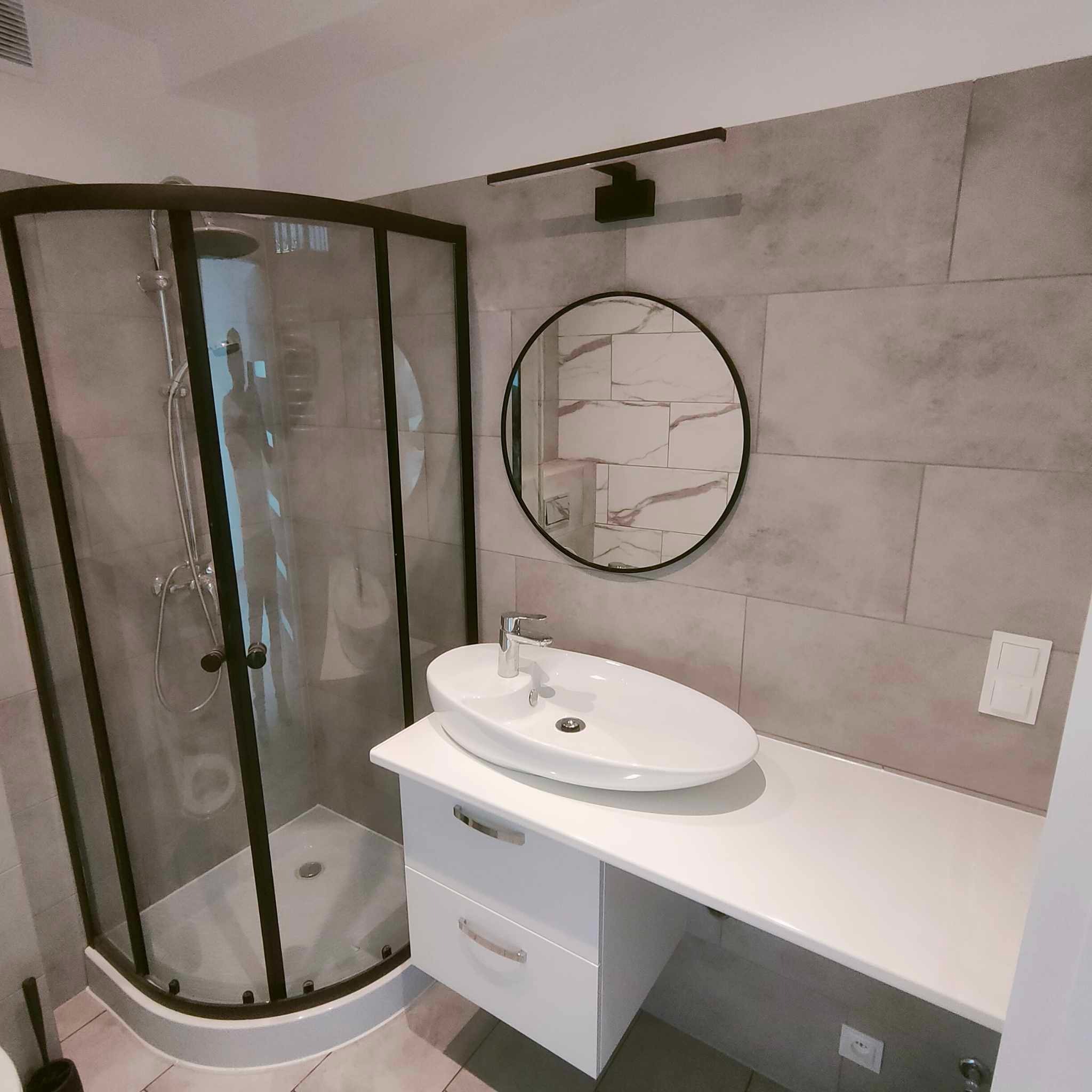 Nowoczesna łazienka z kabiną prysznicową z czarną ramą, owalną umywalką na białej szafce z szufladami i okrągłym lustrem na szarej ścianie wyłożonej płytkami imitującymi kamień.