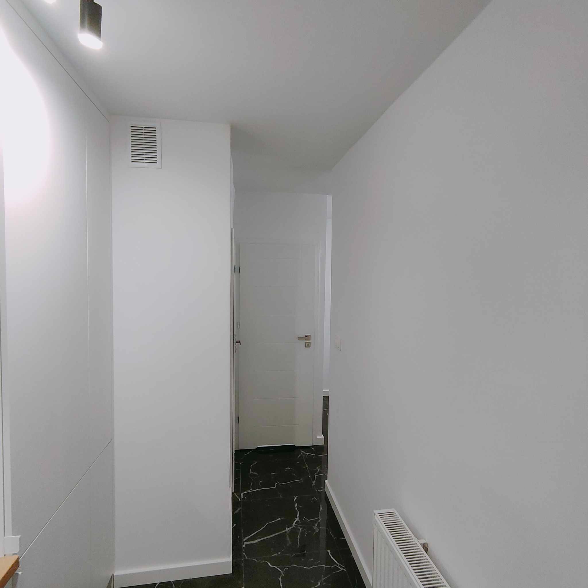 Korytarz z czarną marmurową podłogą, białe ściany z grzejnikiem i wbudowaną szafą, oświetlenie punktowe na suficie, białe drzwi w głębi.