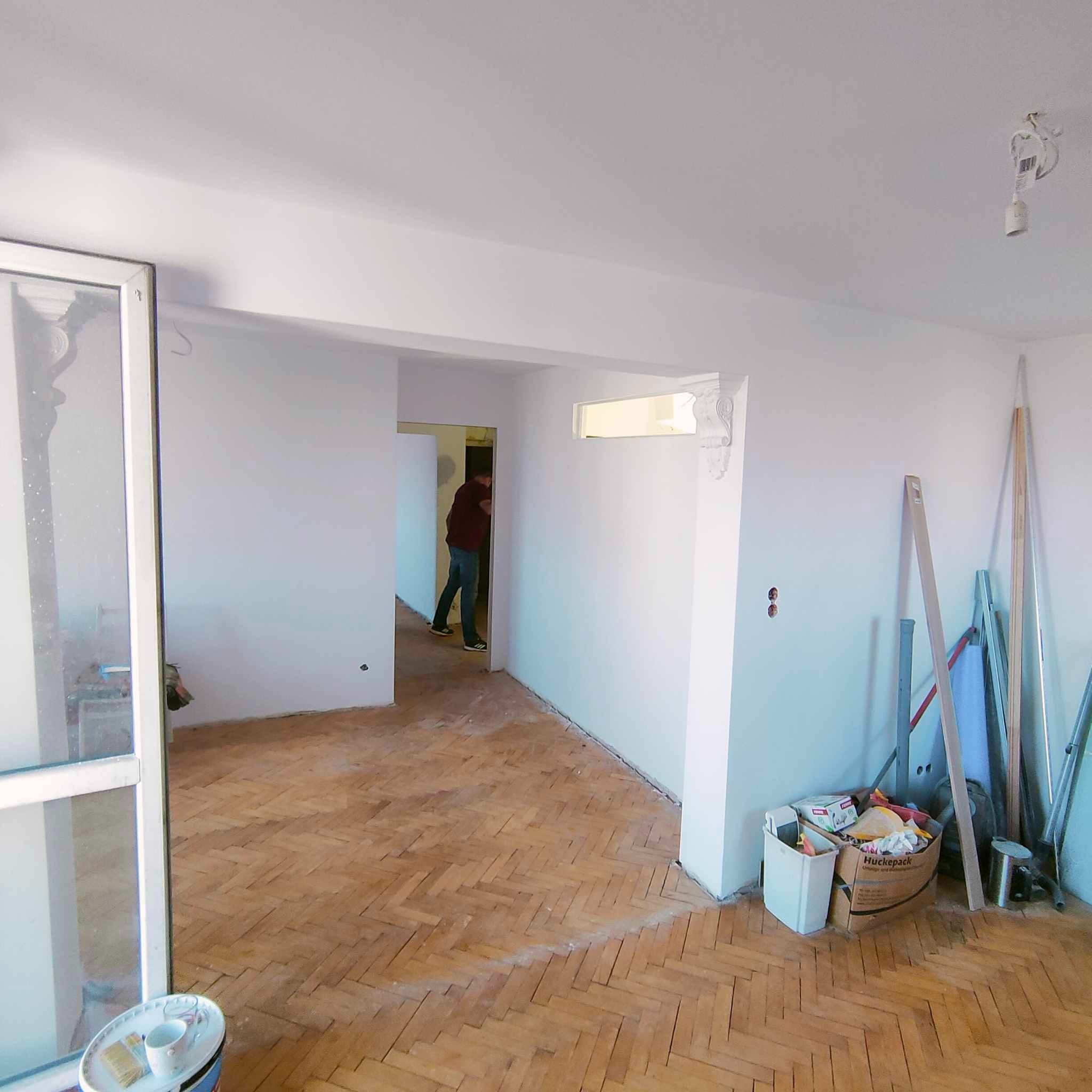 Pokój po malowaniu ścian na biało, z parkietem w jodełkę, widoczne ślady po remoncie, puszka z farbą i kubek na podłodze, w tle osoba przechodząca do kolejnego pomieszczenia.