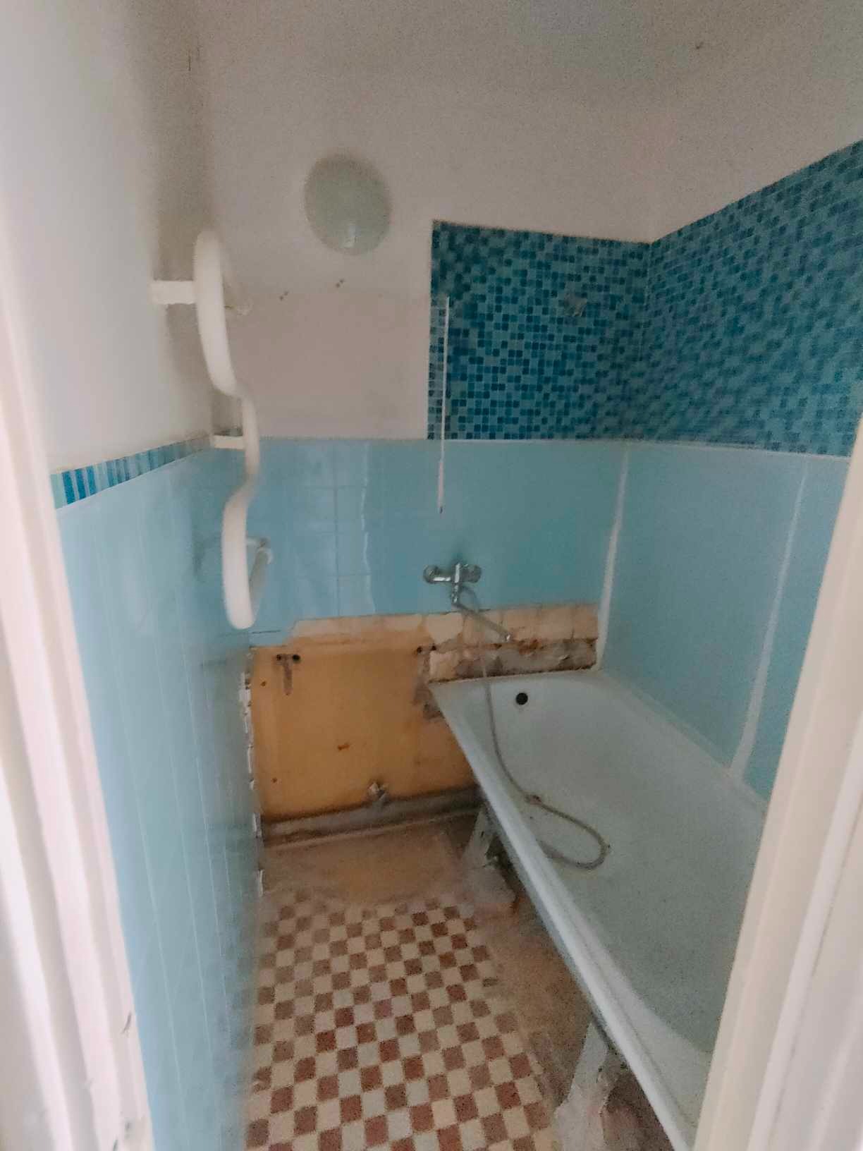 Łazienka w trakcie remontu: częściowo usunięte niebieskie płytki, odsłonięta żółta ściana pod spodem, widoczna wanna i fragment podłogi w szachownicę.