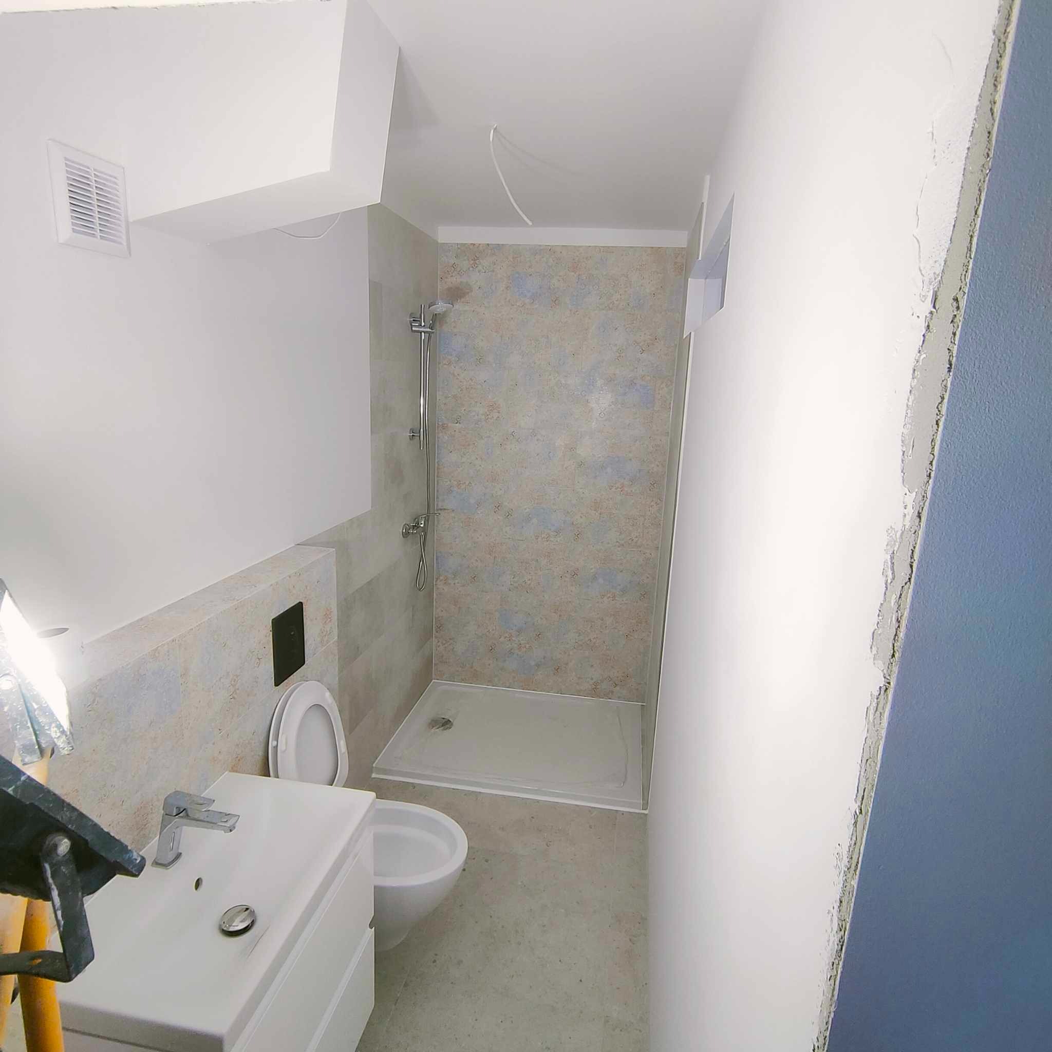 Wnętrze łazienki po remoncie: biała umywalka z szafką, toaleta podwieszana, kabina prysznicowa z jasnymi płytkami z wzorem, wentylator w suficie, fragment ściany z niedokończonymi gładziami.