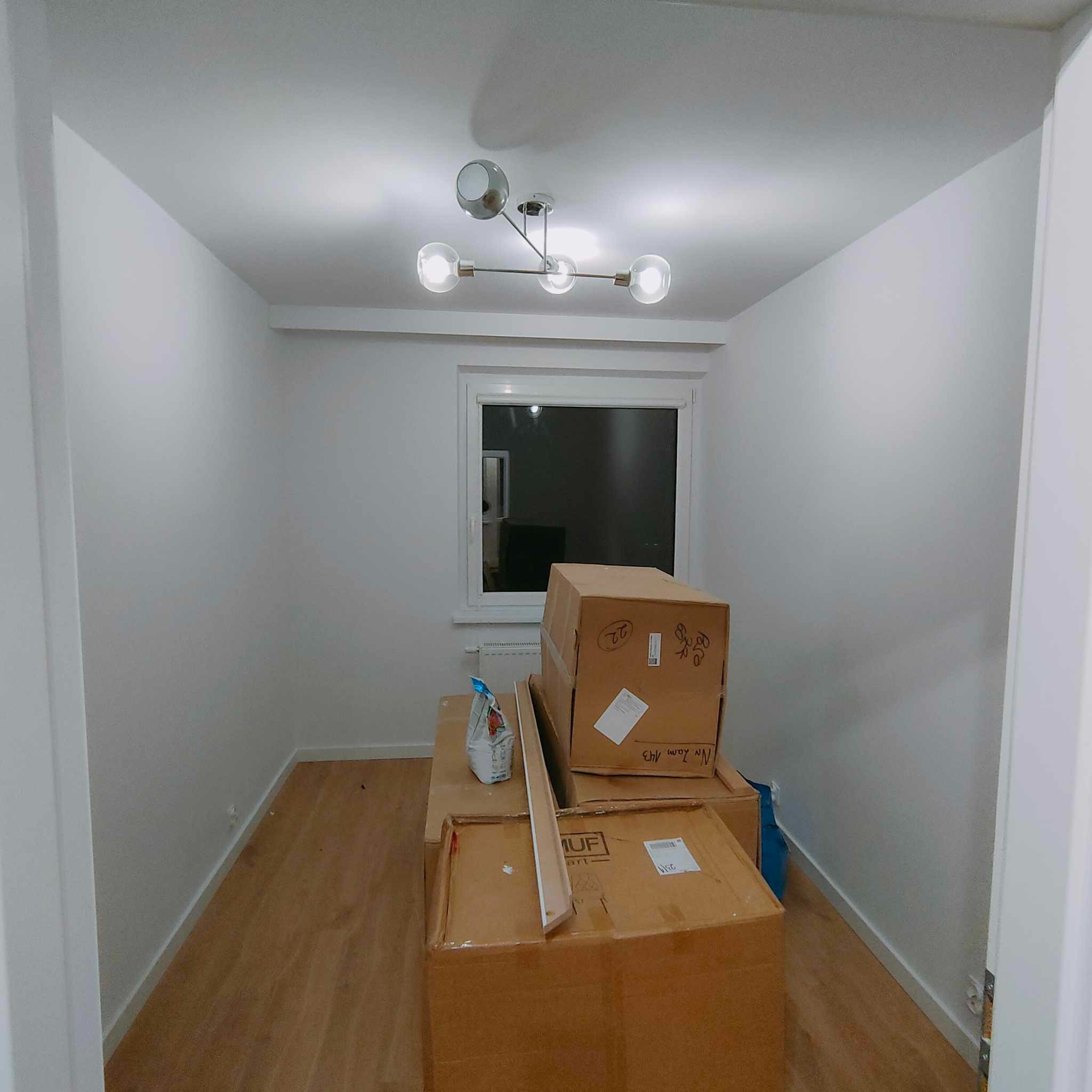 Pusty pokój z drewnianą podłogą i białymi ścianami, oświetlony nowoczesną lampą sufitową, z kartonami po przeprowadzce ustawionymi pod oknem.