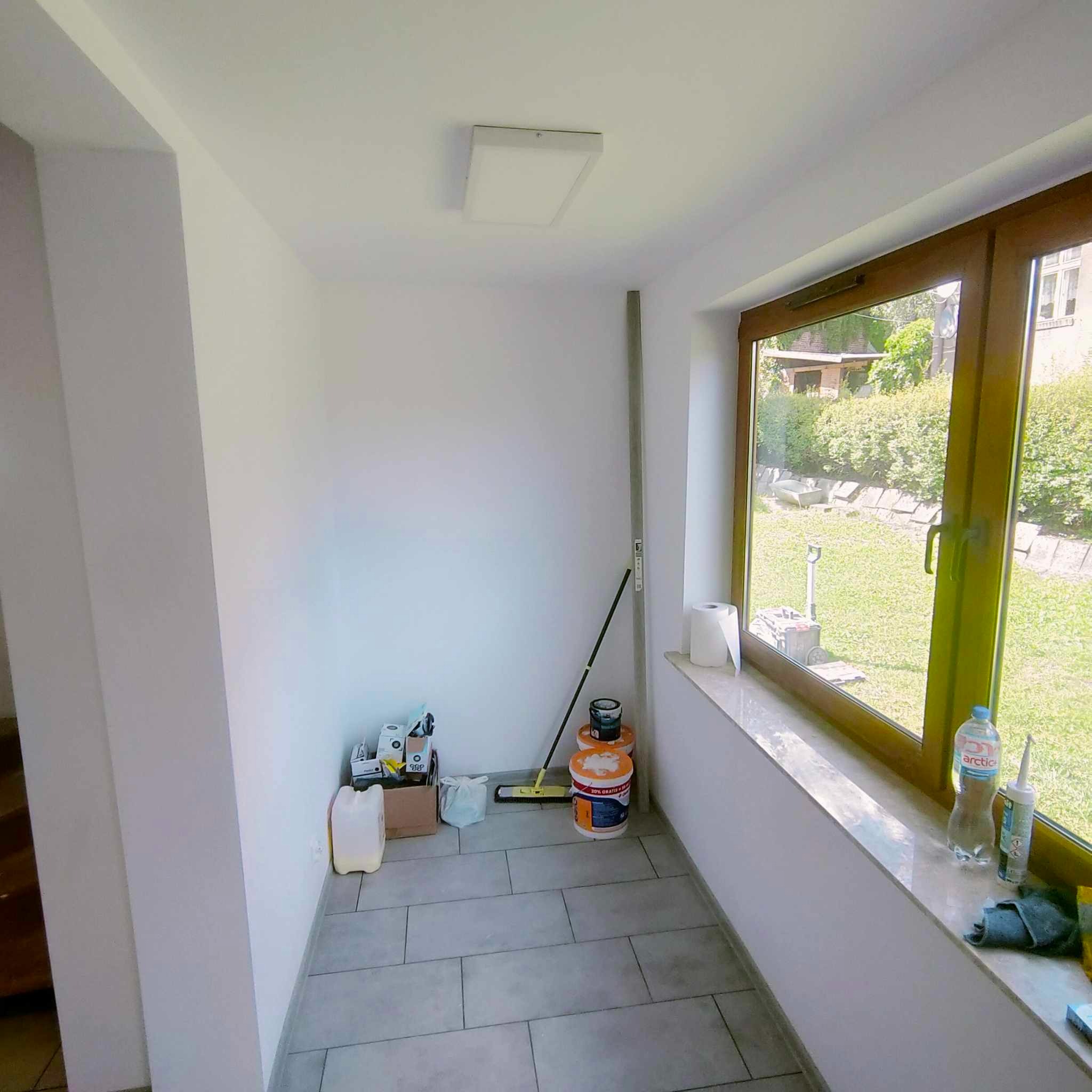 Wnętrze pomieszczenia po malowaniu ścian na biało, widoczne narzędzia malarskie, drabina aluminiowa i puszki z farbą w rogu, okno z widokiem na ogród.
