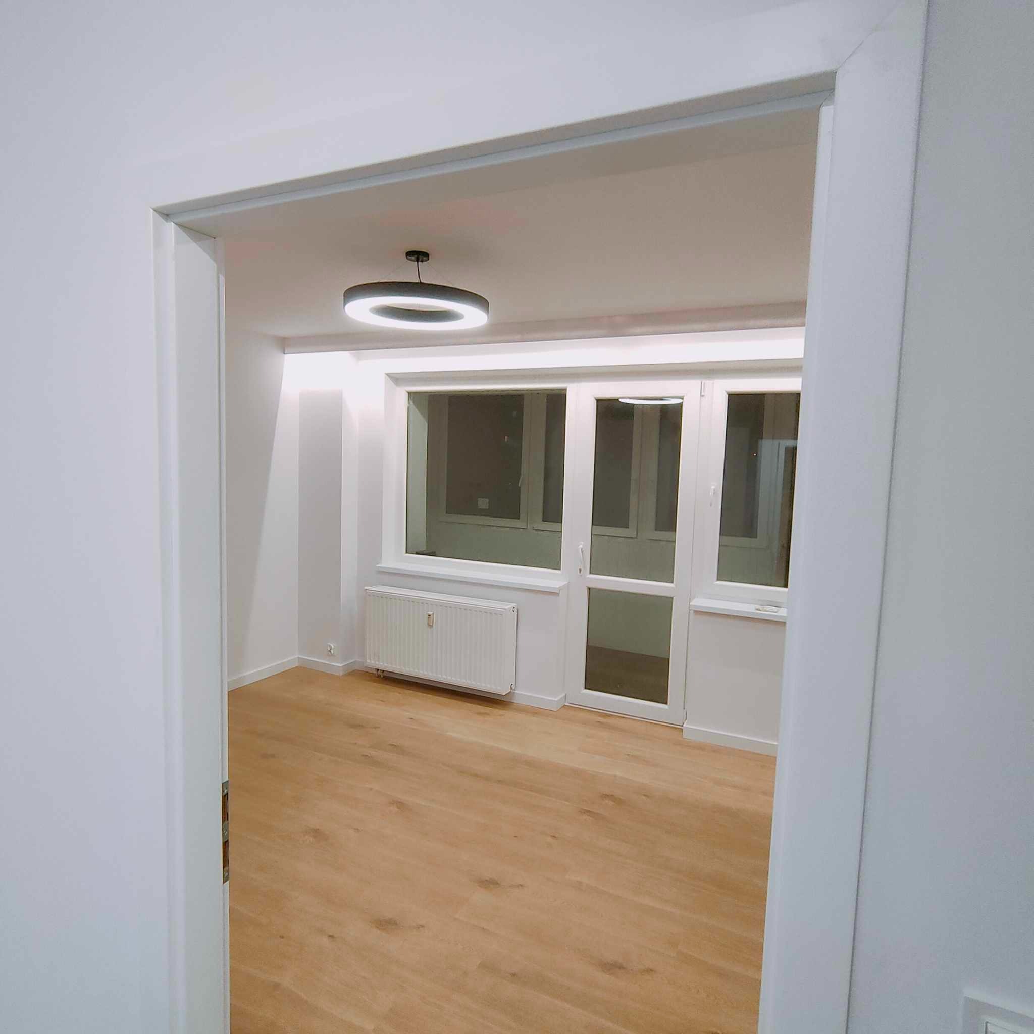 Pusty, jasny pokój po remoncie z drewnianą podłogą, białymi ścianami i nowoczesną okrągłą lampą sufitową, widok zza futryny drzwi.