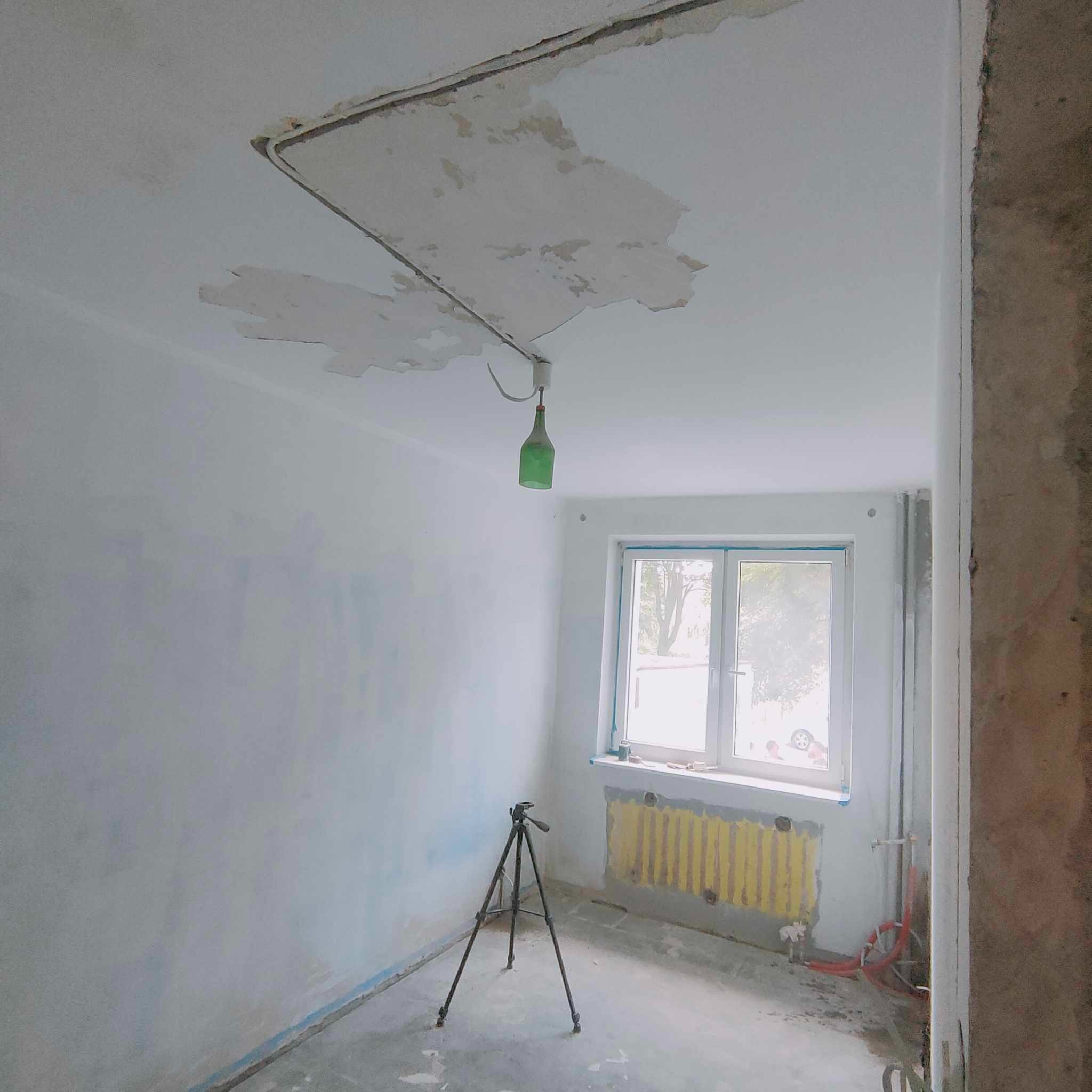 Pokój w trakcie przygotowania do malowania: częściowo zdarta stara farba z sufitu, widoczne ślady po instalacji elektrycznej, żółty grzejnik pod oknem, statyw fotograficzny na podłodze.