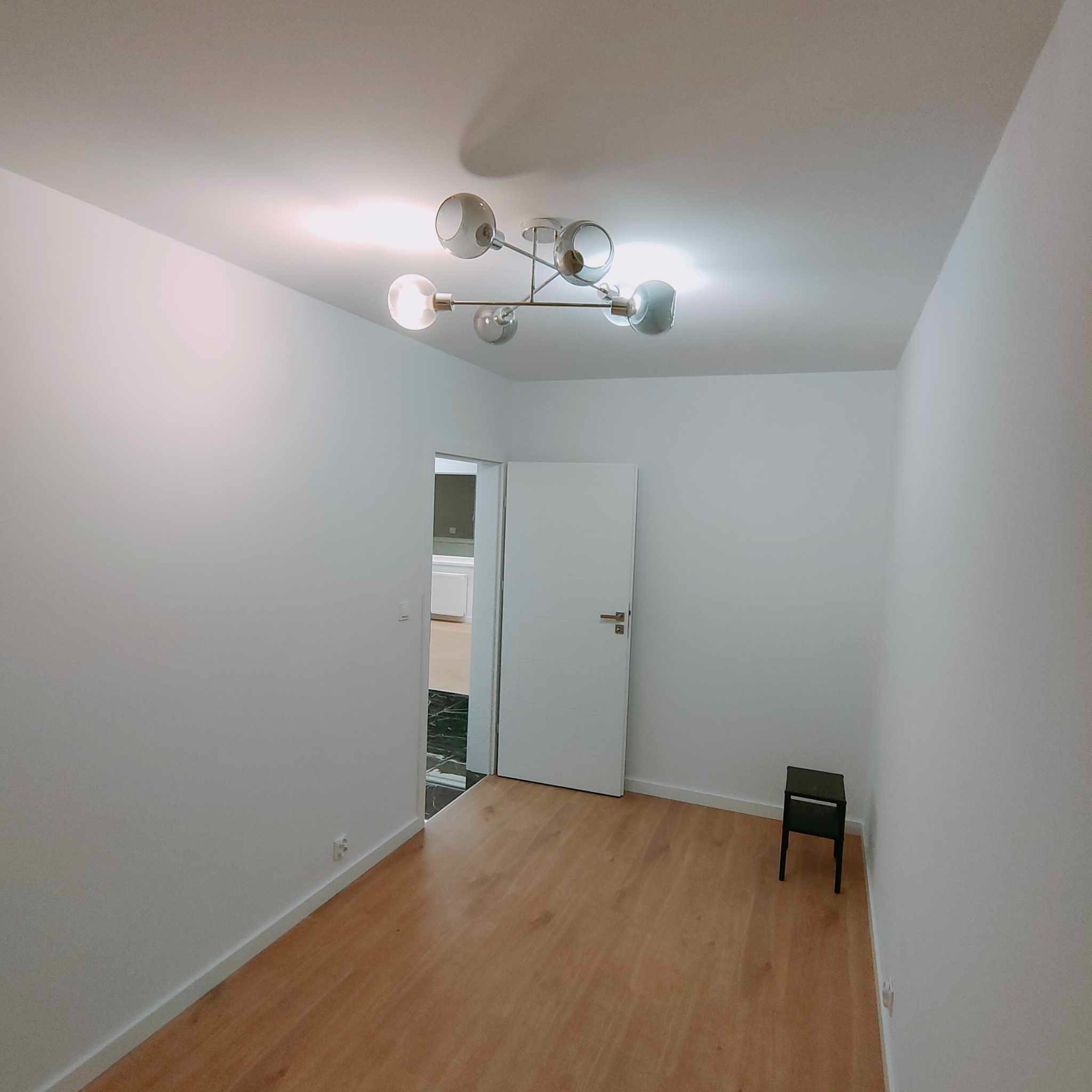 Pusty pokój z drewnianą podłogą, białymi ścianami i designerską lampą sufitową z kilkoma szklanymi kloszami, widok na otwarte drzwi do kolejnego pomieszczenia z czarną podłogą i białymi meblami.