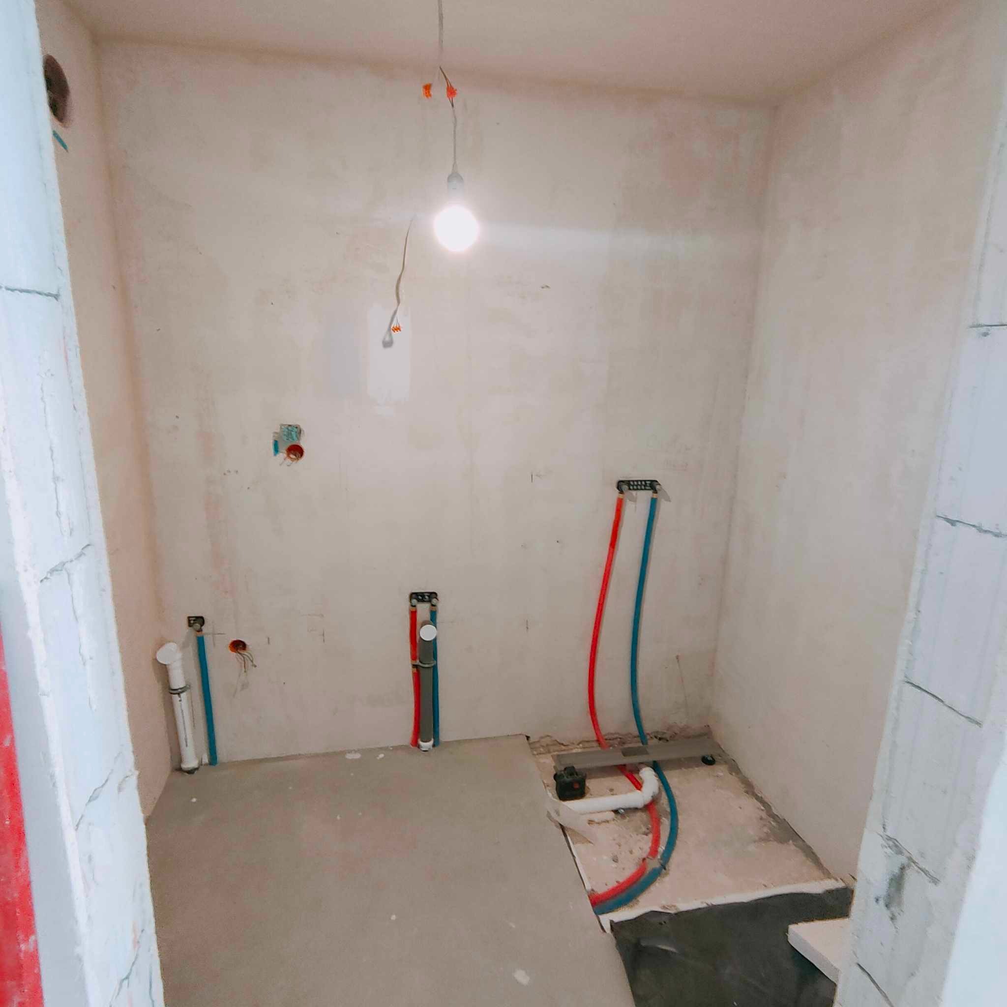 Surowa łazienka w trakcie prac instalacyjnych: widoczne rury wodne w kolorze czerwonym i niebieskim, wystające ze ściany kable elektryczne oraz surowe ściany przygotowane do dalszego wykończenia.