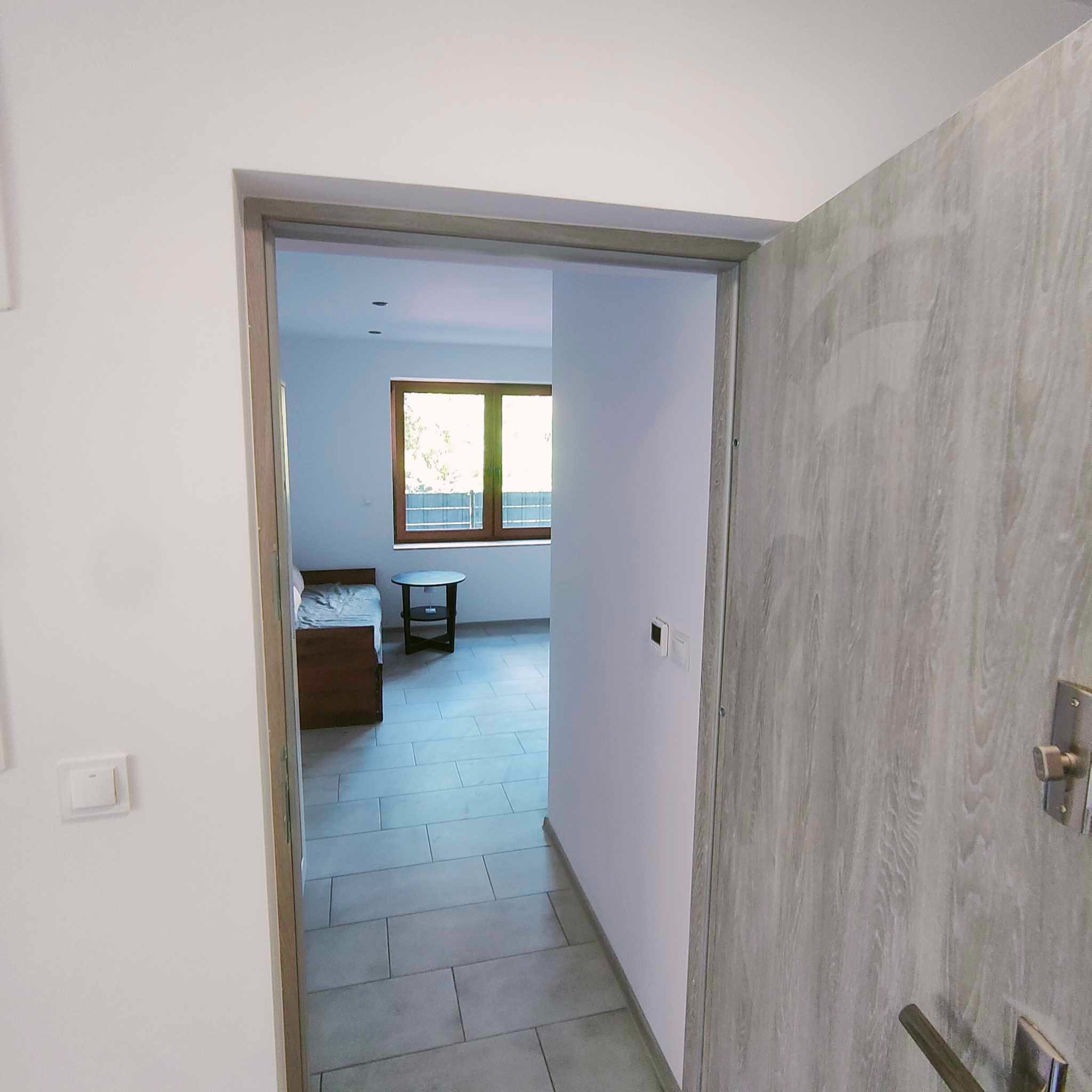 Widok przez otwarte drzwi do pokoju z szarymi płytkami podłogowymi, drewnianym łóżkiem i stolikiem przy oknie.