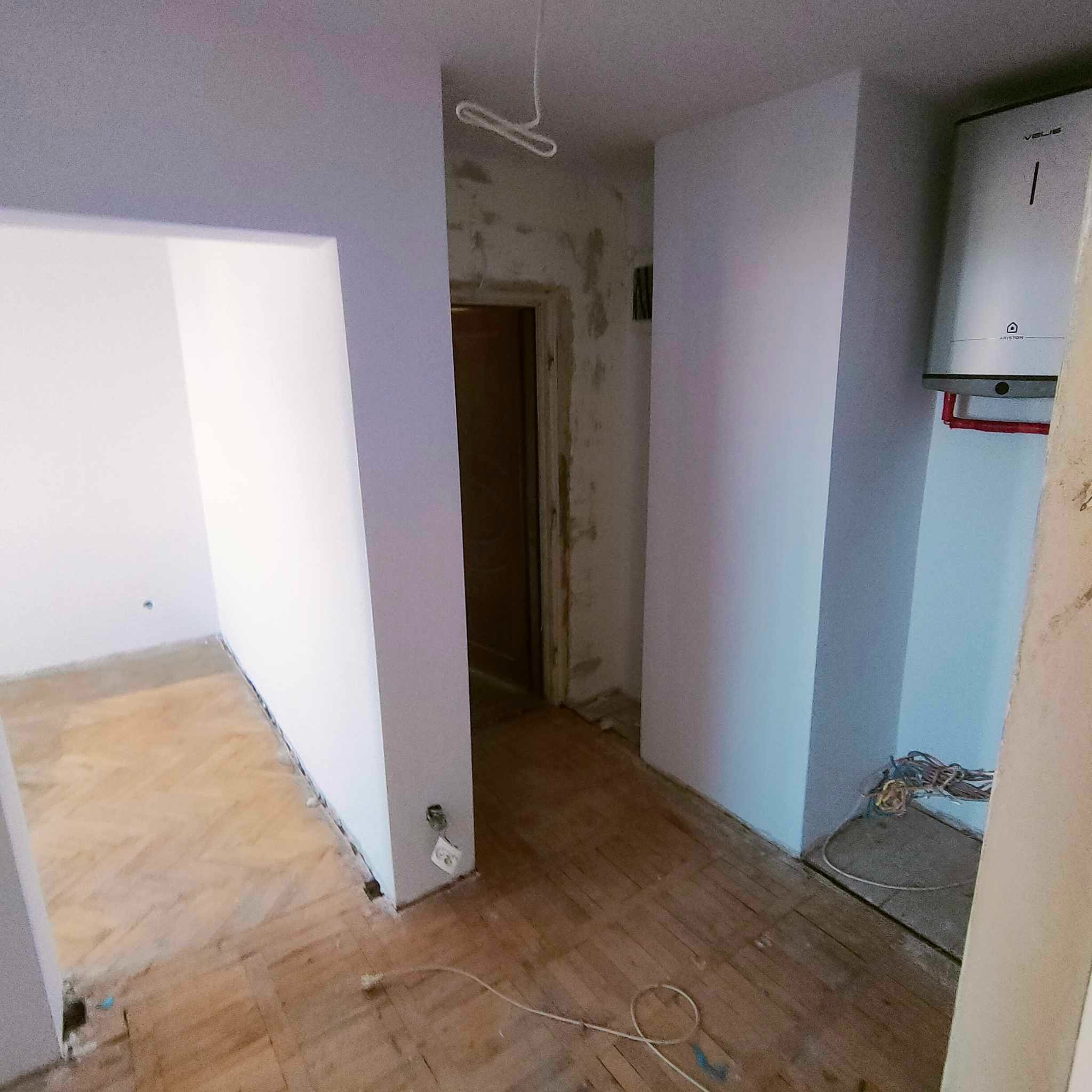 Wnętrze mieszkania w trakcie remontu: świeżo pomalowane ściany na szaro, drewniana podłoga, widoczne kable elektryczne, bojler na ścianie i niedokończone wykończenie futryny drzwiowej.