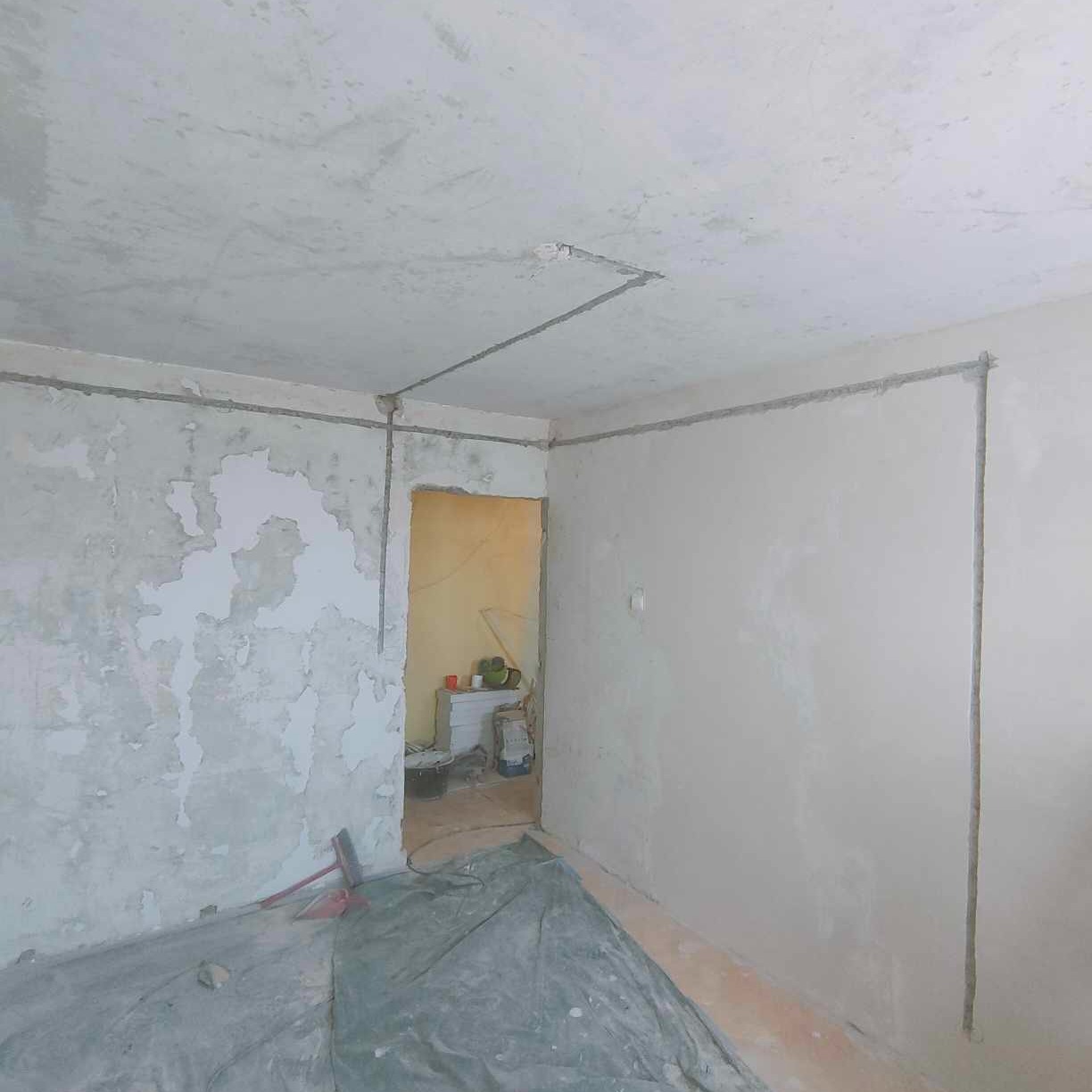 Pokój w trakcie remontu: ściana z częściowo usuniętą starą farbą, nowa gładź na ścianie obok, zabezpieczona podłoga, widoczne przewody elektryczne prowadzone po ścianach i suficie.