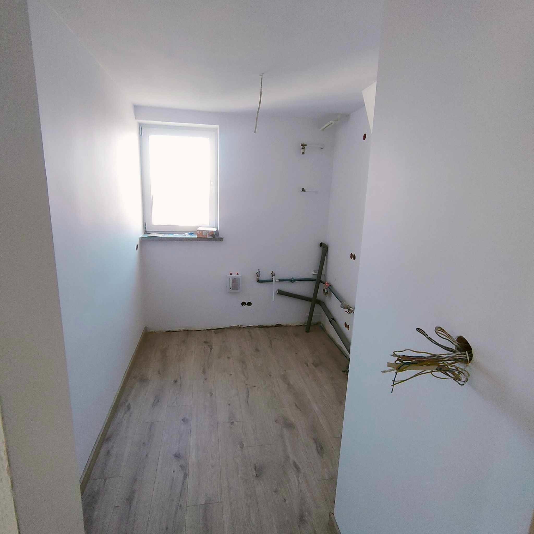 Surowe wnętrze łazienki w trakcie prac: widoczne rury instalacji wodnej, wystające przewody elektryczne, jasne ściany i podłoga imitująca drewno, okno z parapetem.