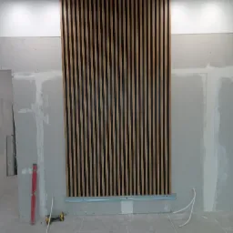 Montaż drewnianej ażurowej ścianki dekoracyjnej na metalowym stelażu, widoczna poziomica i wiertarka akumulatorowa, ściana w trakcie wykańczania płytami gipsowo-kartonowymi.