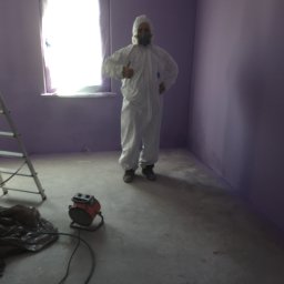 Grzegorz Debski - Pracownik w kombinezonie ochronnym i masce daje kciuk w górę w świeżo pomalowanym na fioletowo pokoju, widoczna drabina i nagrzewnica elektryczna.