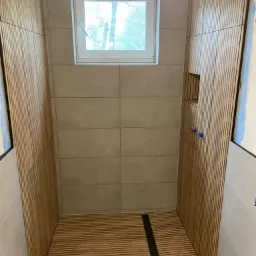 Nowoczesna kabina prysznicowa z drewnianymi panelami na ścianach i podłodze, odpływ liniowy, okno z widokiem na drzewa, częściowo wykończona.