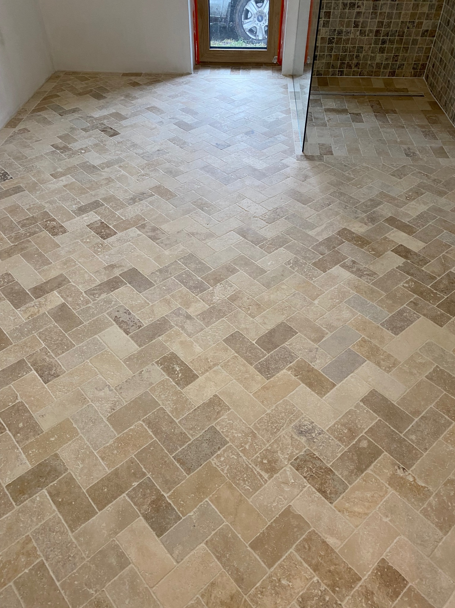 Beżowa podłoga z płytek ułożonych w jodełkę w łazience z kabiną prysznicową w rogu i oknem z widokiem na oponę samochodu.