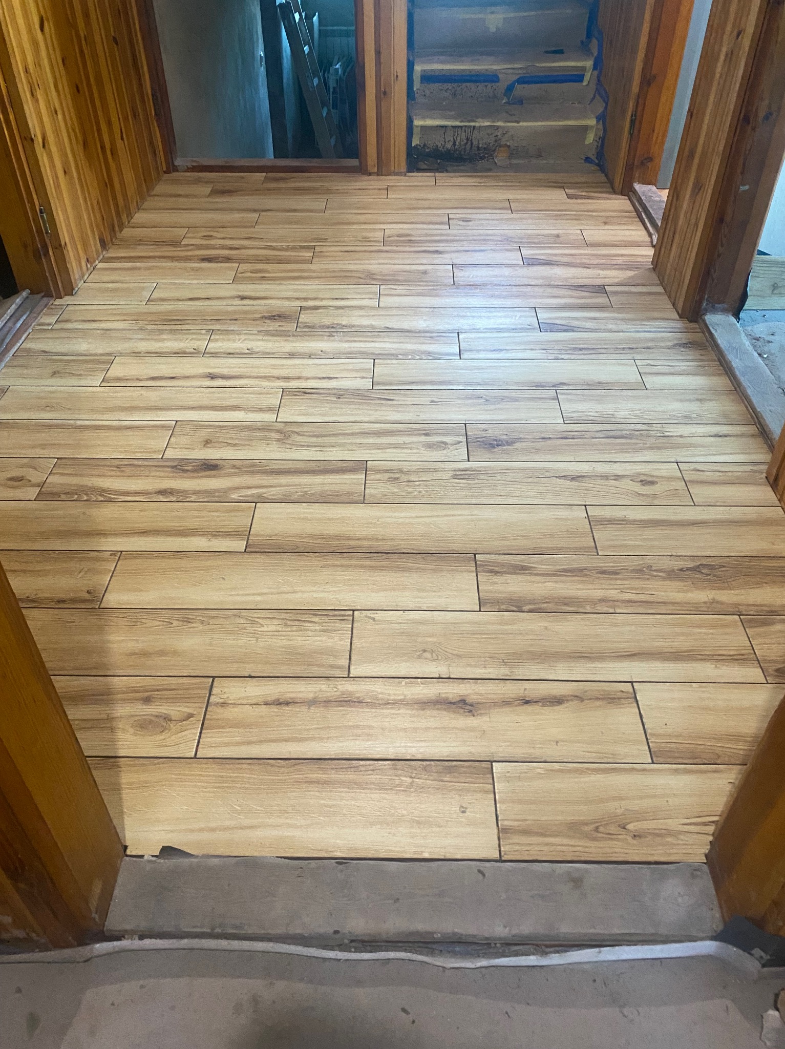 Ułożona podłoga z płytek imitujących drewno, widok z progu pomieszczenia z drewnianymi futrynami, w tle schody w trakcie zabezpieczania folią malarską.