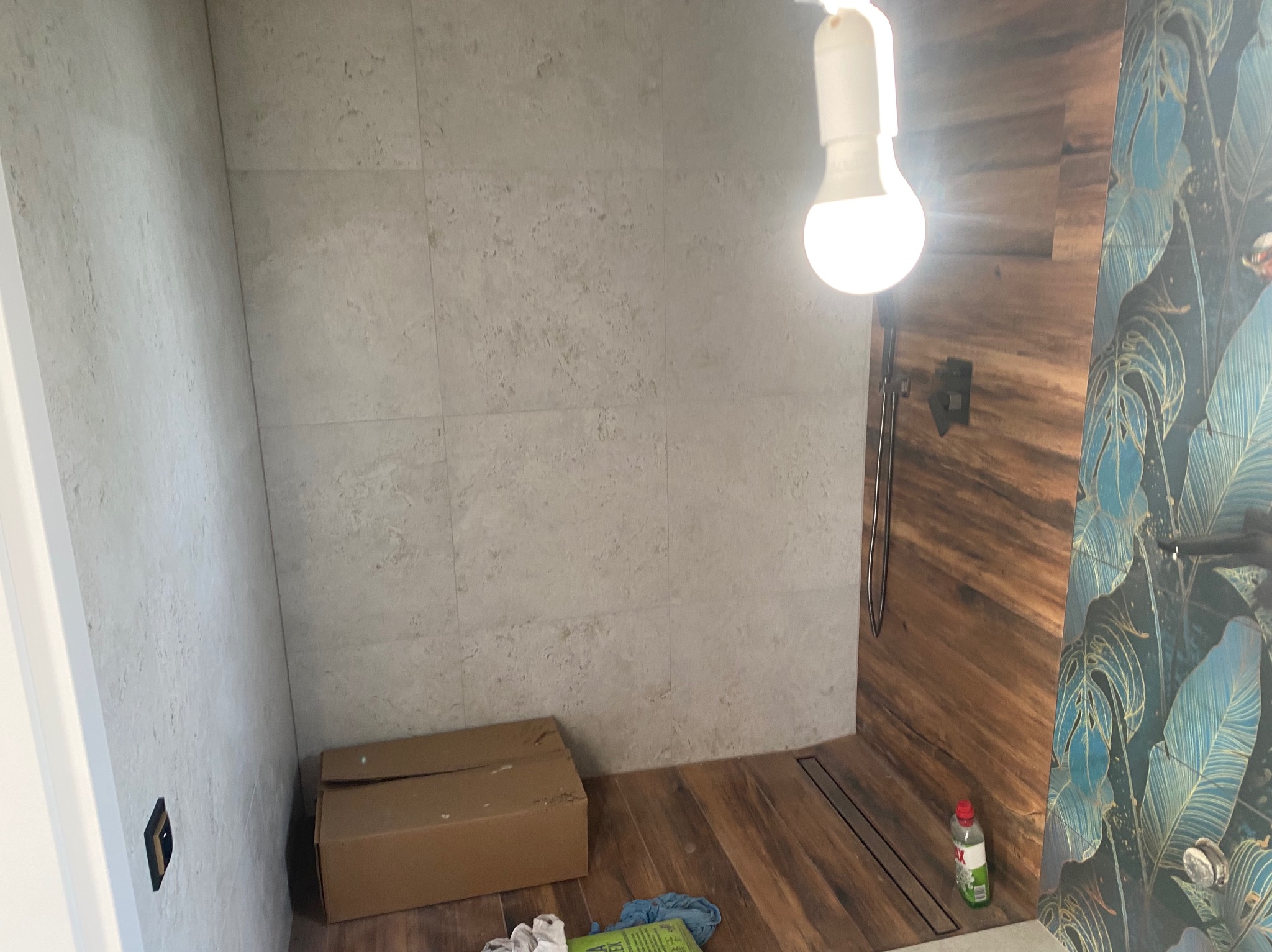 Wnętrze łazienki w trakcie prac wykończeniowych: szare płytki na ścianach, drewnopodobna podłoga, fragment ściany z tapetą w motywy roślinne, pudełko kartonowe i akcesoria na podłodze, surowa...