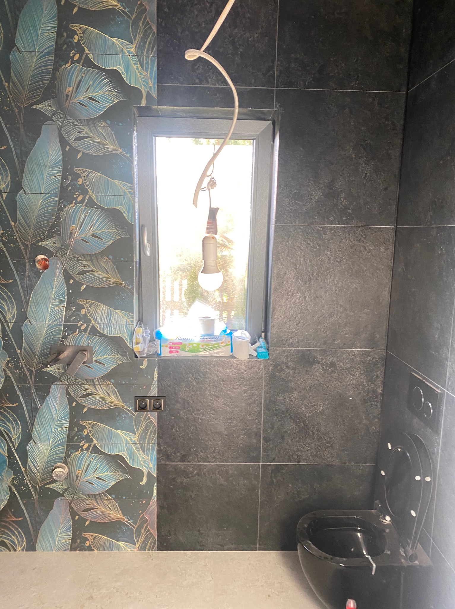 Częściowo wykończona łazienka w Kamieńsku z czarną armaturą i płytkami w roślinne wzory, widoczna wisząca toaleta i okno z akcesoriami.