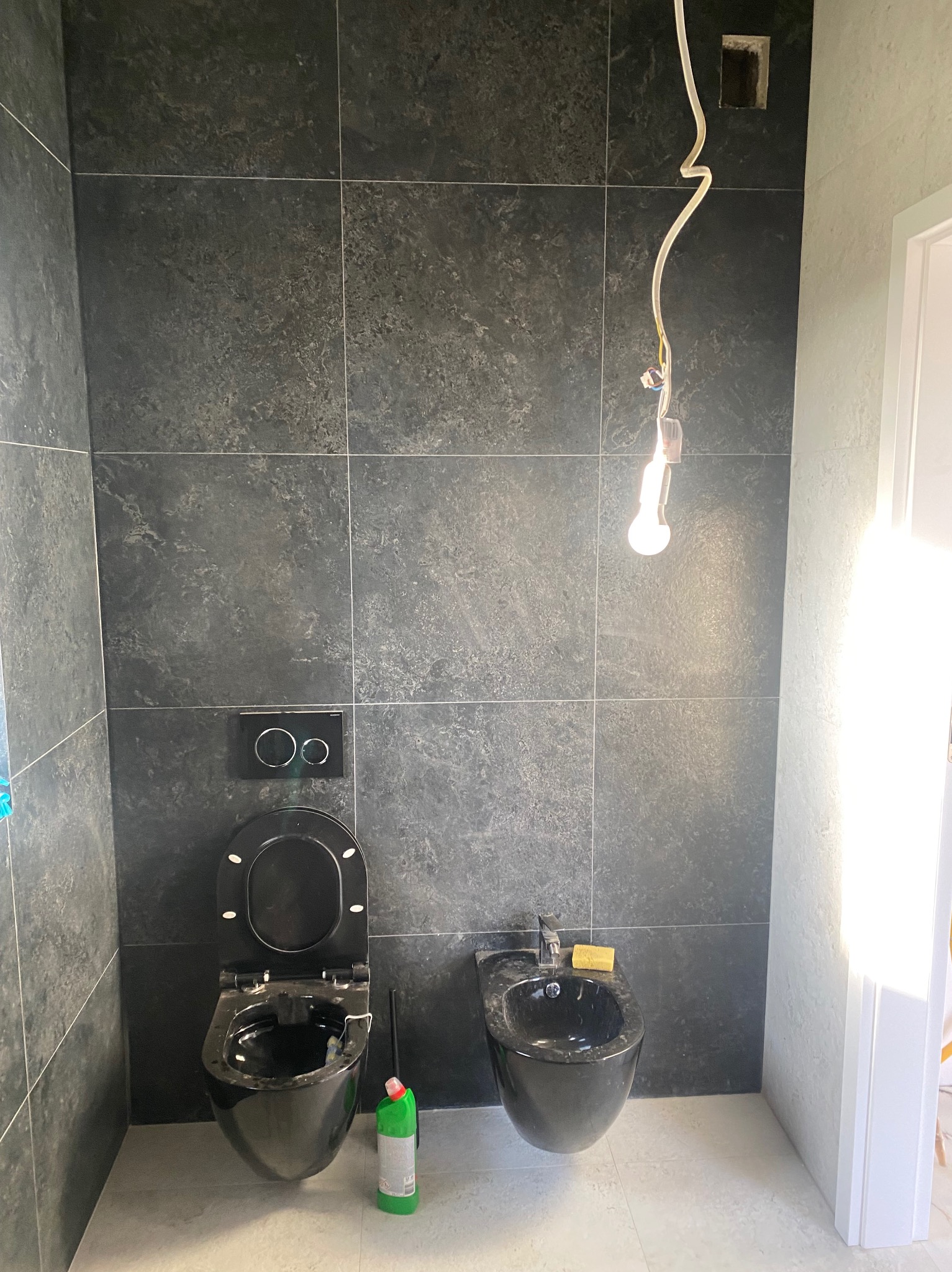 Czarne płytki na ścianach łazienki w trakcie montażu, wisząca toaleta i bidet w kolorze czarnym, surowa żarówka zwisająca z sufitu, butelka środka czyszczącego obok toalety.