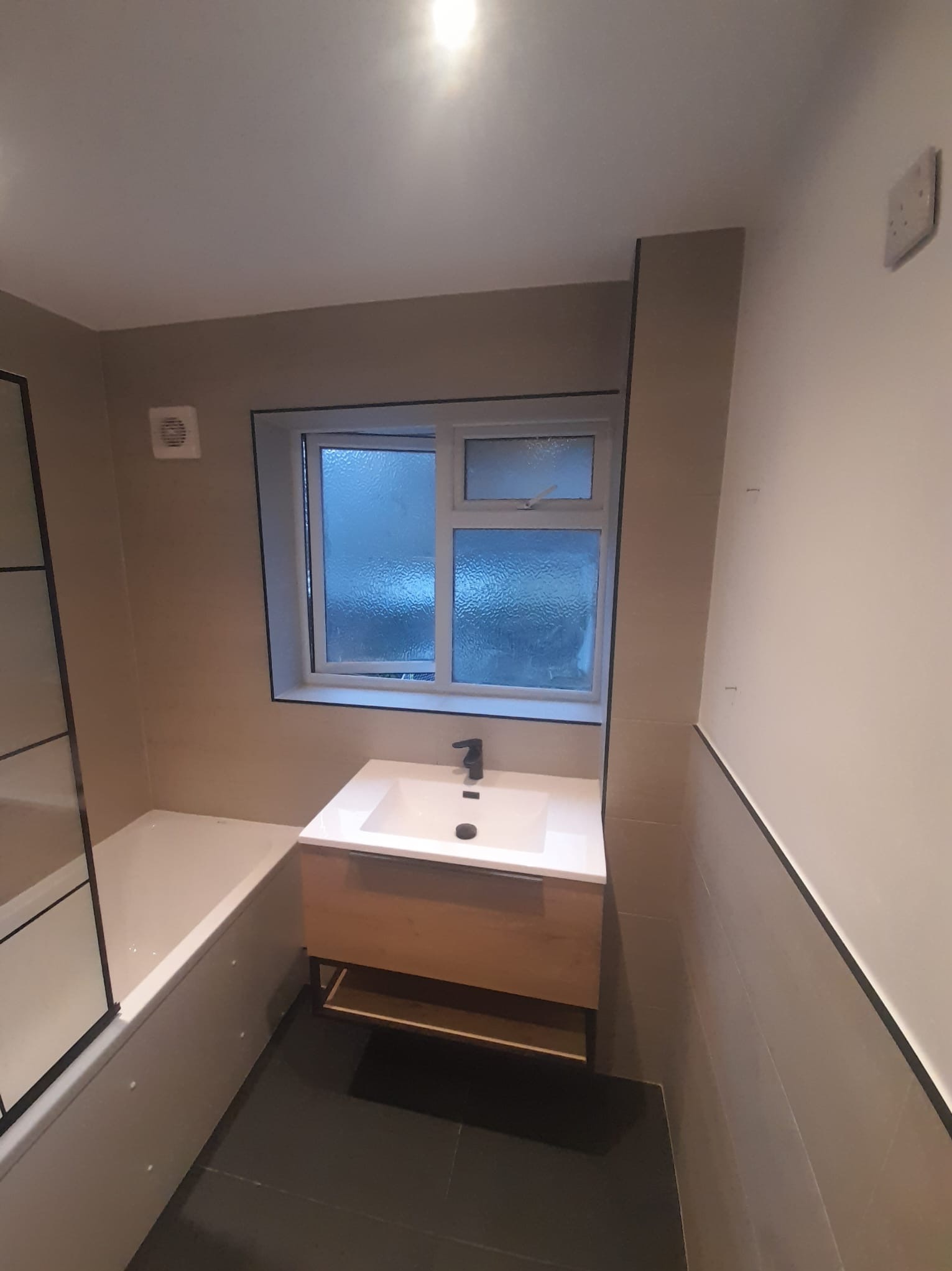 Nowoczesna łazienka z umywalką nablatową na drewnianej szafce, czarną armaturą i czarnymi fugami, widok na okno z zewnątrz, ciemna podłoga.