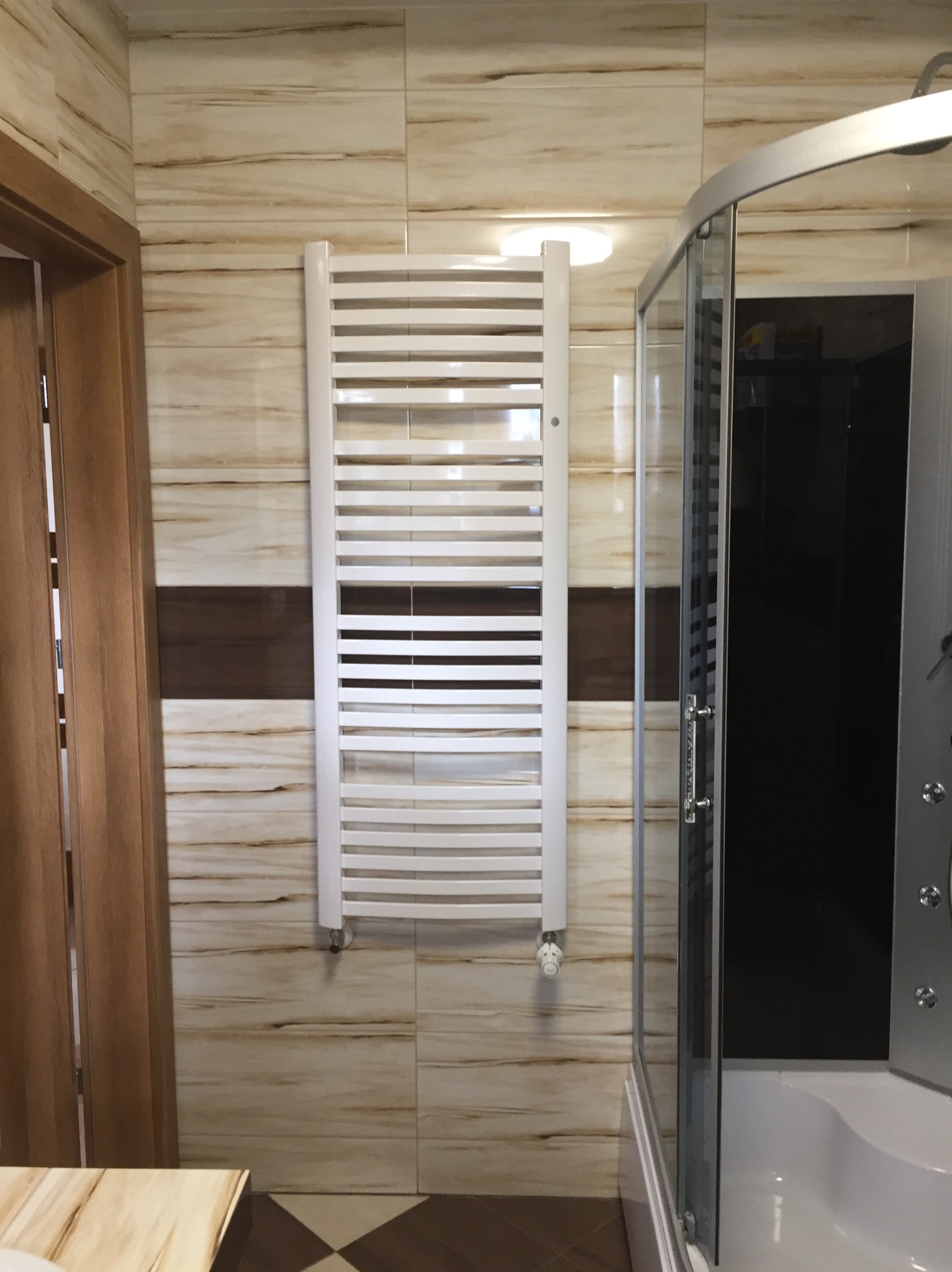 Nowoczesna łazienka z beżowymi płytkami imitującymi drewno, grzejnikiem drabinkowym i kabiną prysznicową z czarnym panelem.