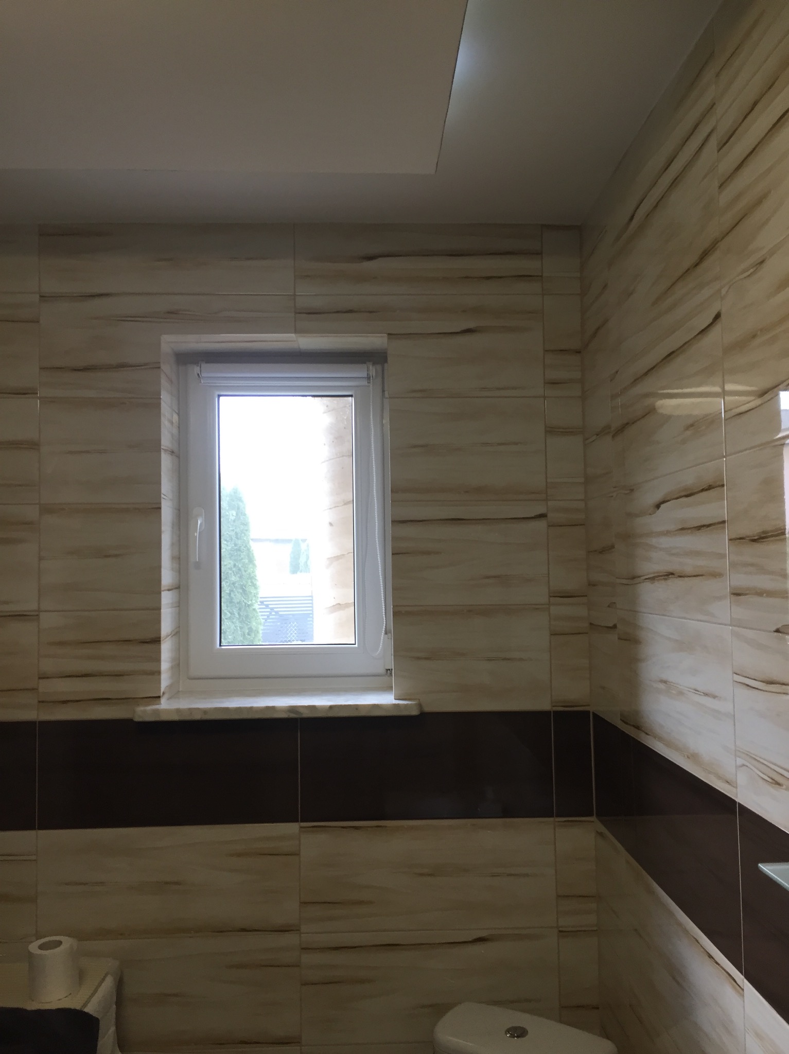 Wykończona łazienka z beżowymi płytkami imitującymi drewno, ciemnobrązowy pas płytek, białe okno z widokiem na zieleń, toaleta w dolnej części kadru.