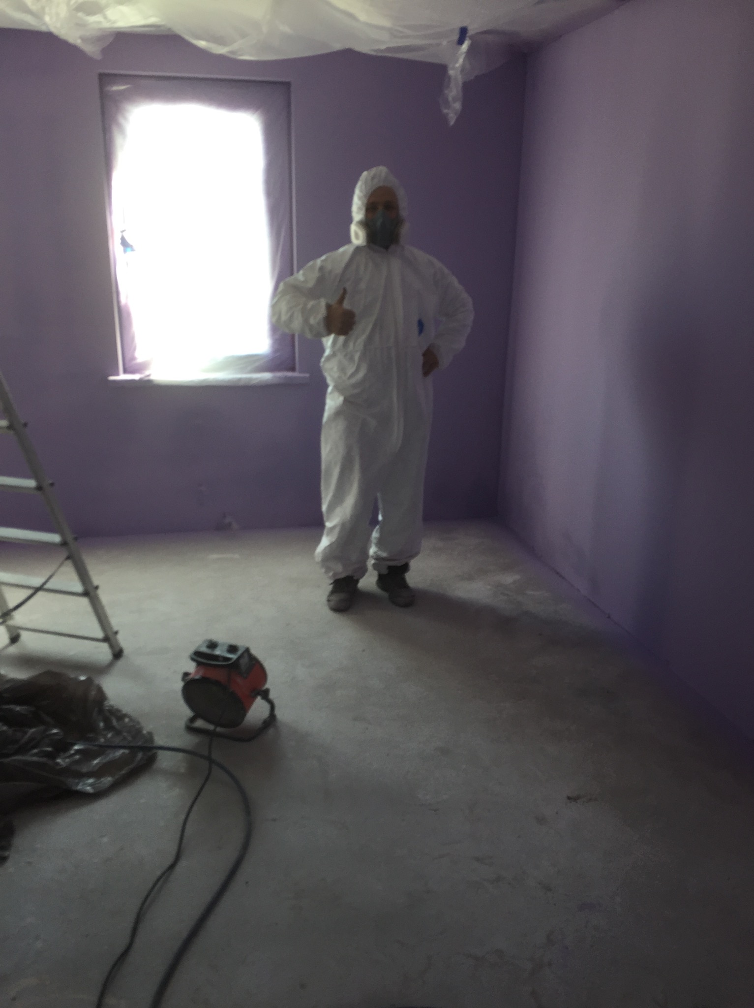 Pracownik w kombinezonie ochronnym i masce daje kciuk w górę w świeżo pomalowanym na fioletowo pokoju, widoczna drabina i nagrzewnica elektryczna.