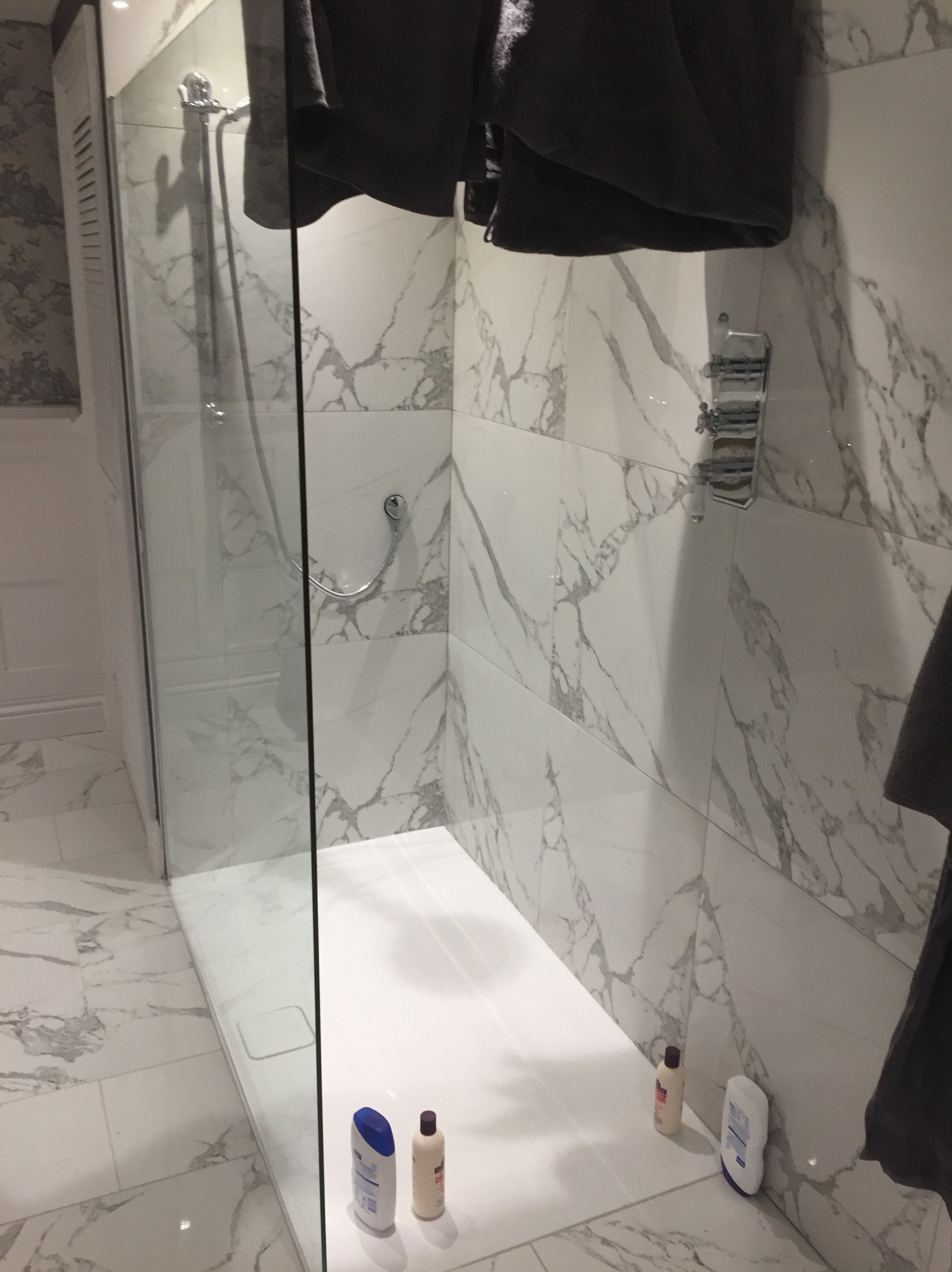 Nowoczesna kabina prysznicowa z marmurowymi płytkami, szklaną ścianą i kilkoma butelkami kosmetyków na brodziku.