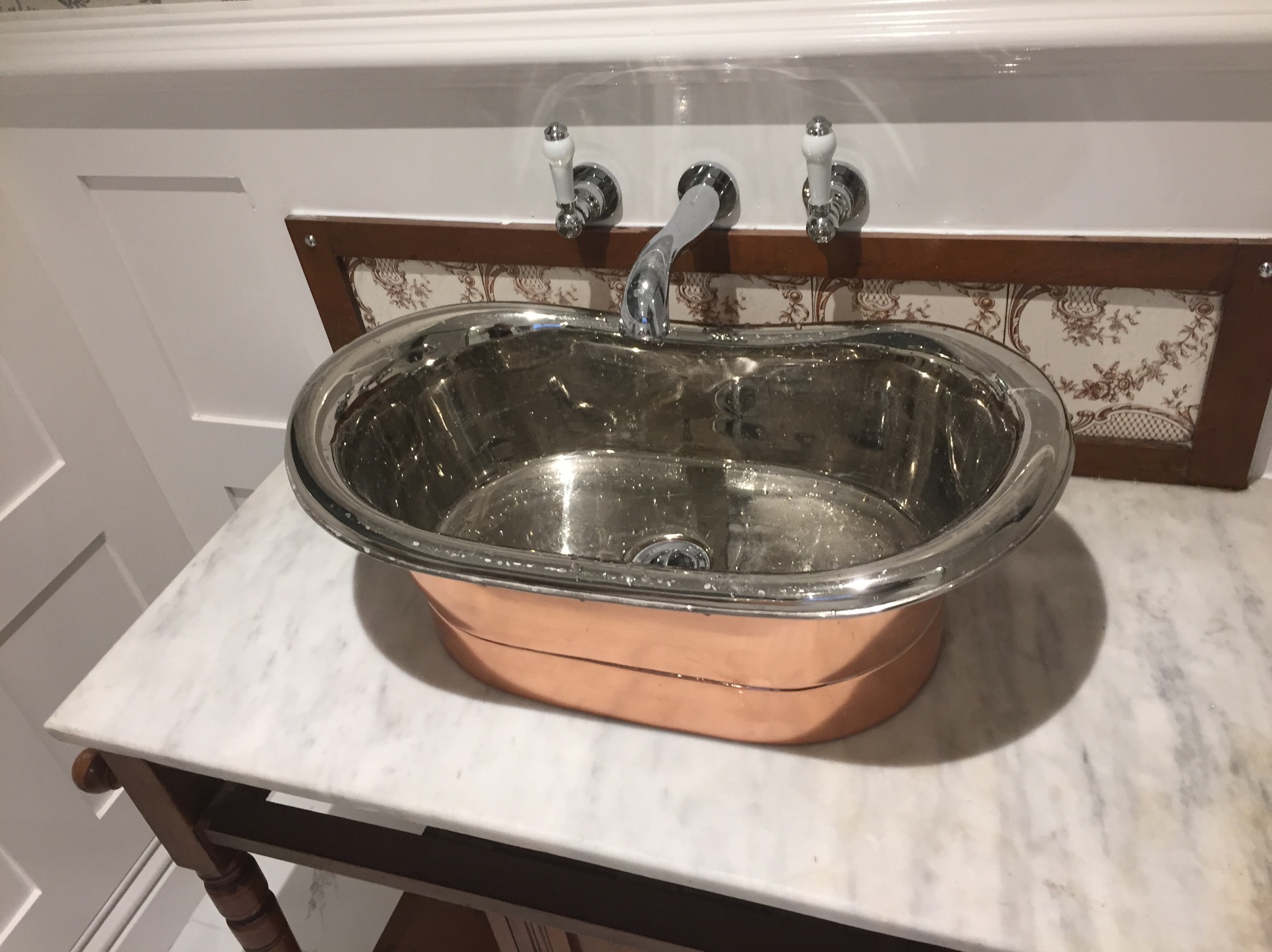 Miedziano-srebrna umywalka w stylu retro na marmurowym blacie, z baterią z porcelanowymi uchwytami i ozdobnym panelem ściennym.