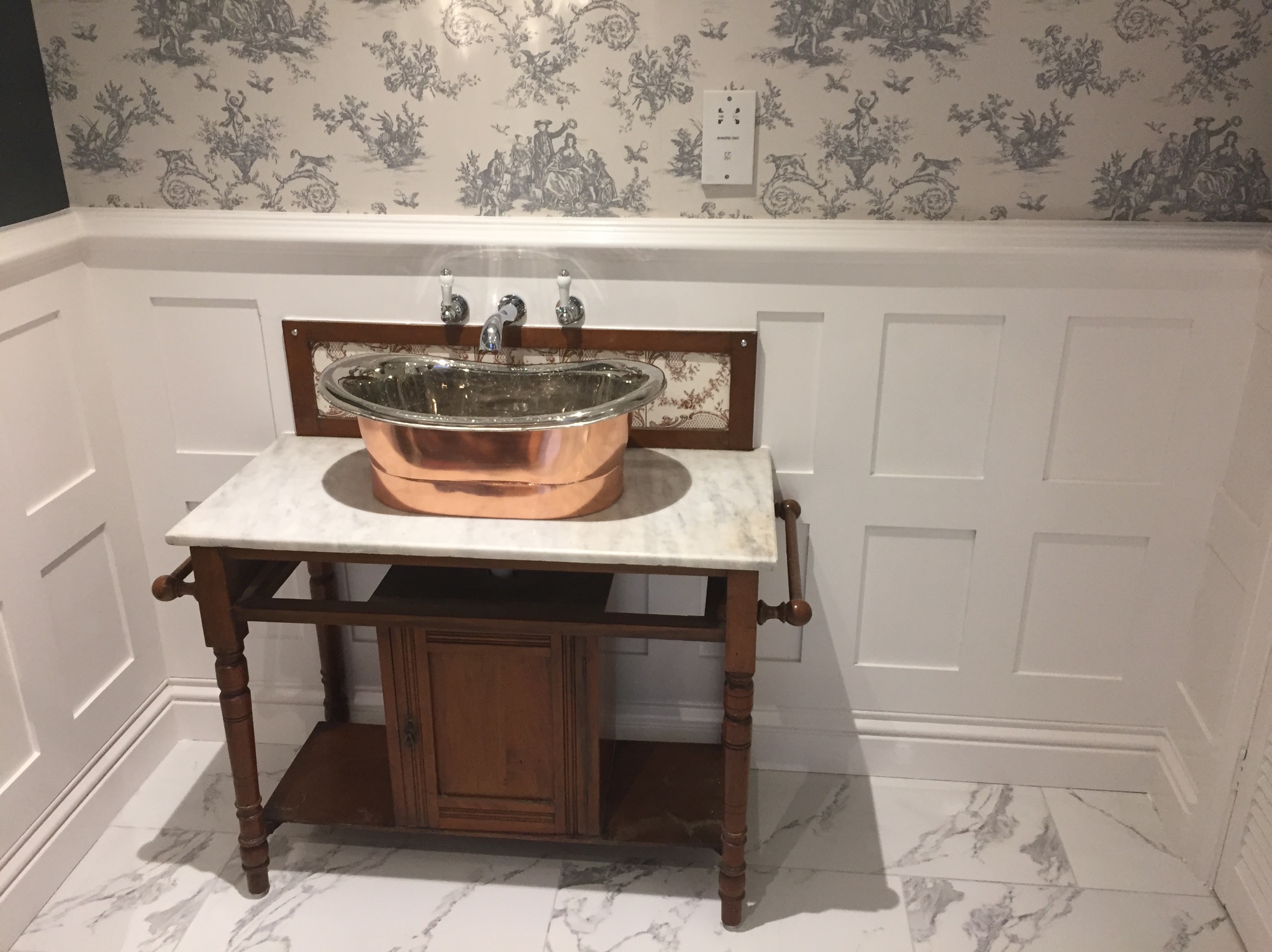 Miedziana umywalka na drewnianym postumencie z marmurowym blatem, zamontowana w łazience z białą boazerią i tapetą w stylu toile de jouy.