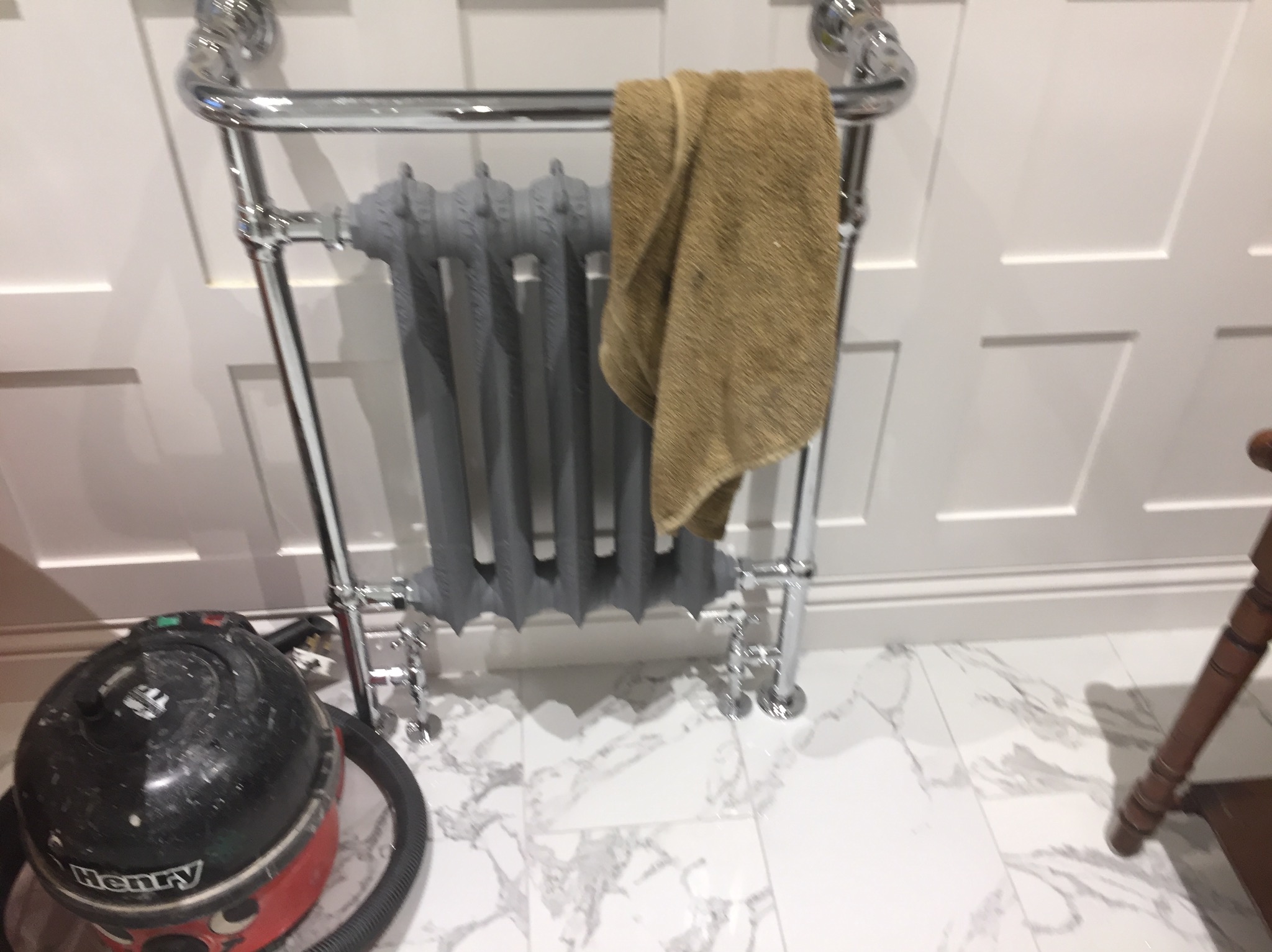 Szary, żeliwny grzejnik łazienkowy z chromowanymi rurami i ręcznikiem, umieszczony na marmurowej podłodze, obok odkurzacza Henry.