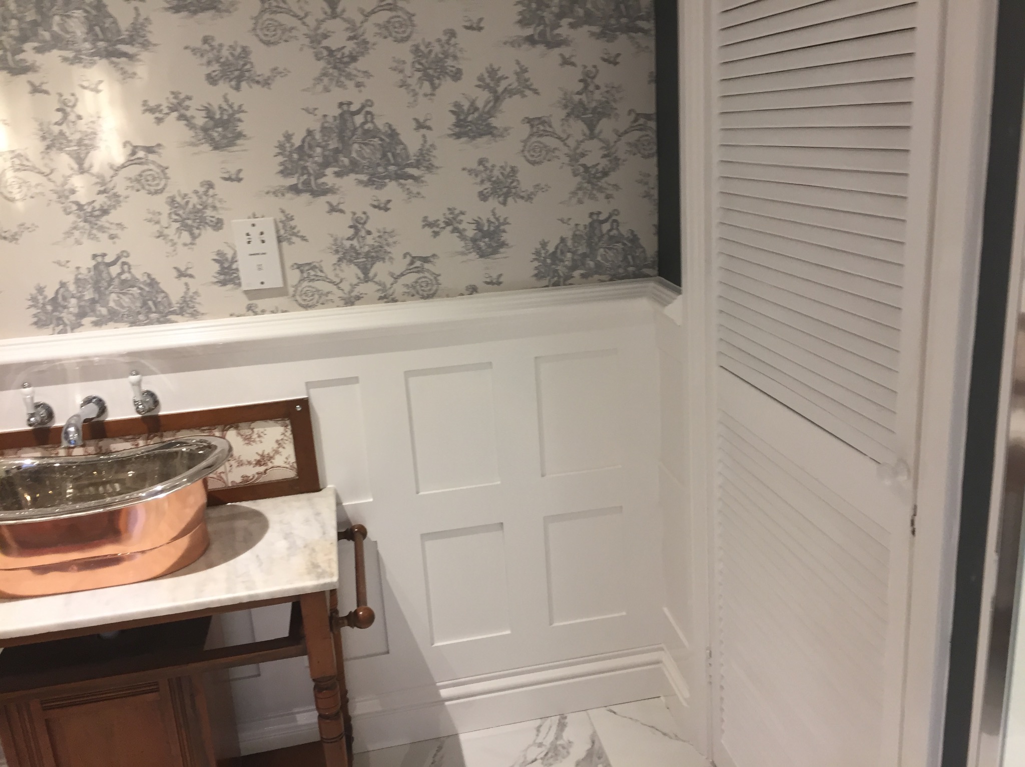 Stylowa łazienka z miedzianą umywalką na marmurowym blacie, wzorzystą tapetą w stylu toile de jouy i białymi panelami ściennymi.