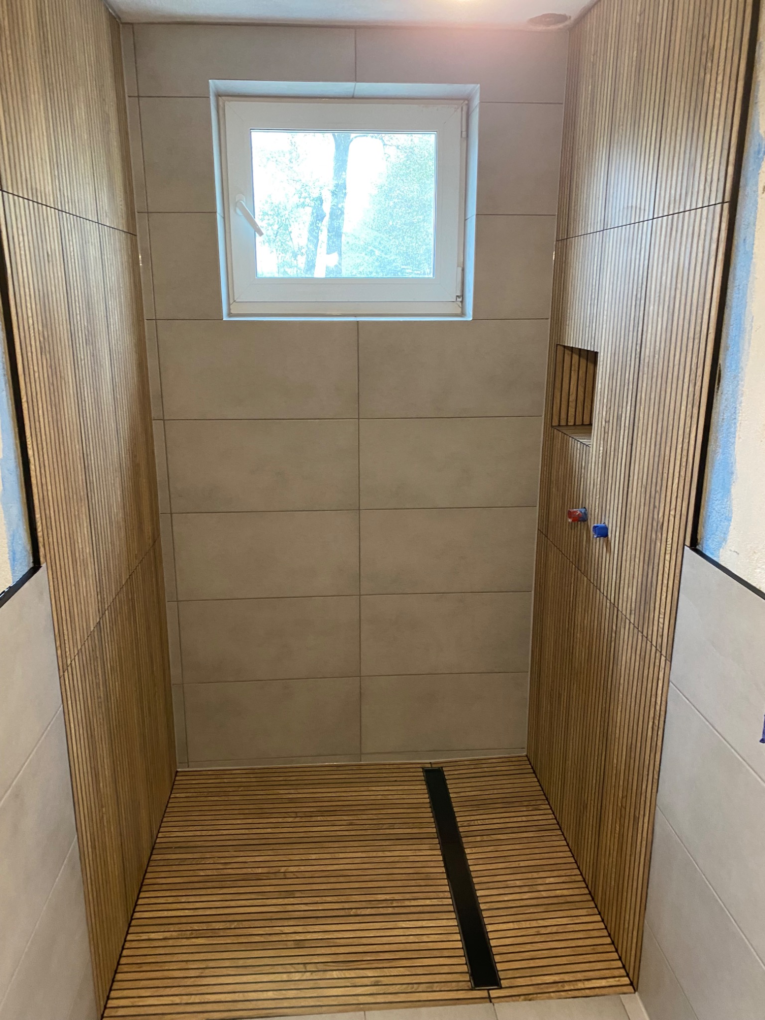 Nowoczesna kabina prysznicowa z drewnianymi panelami na ścianach i podłodze, odpływ liniowy, okno z widokiem na drzewa, częściowo wykończona.