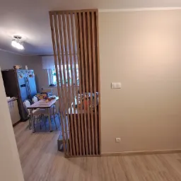 Drewniana ażurowa ścianka działowa oddzielająca kuchnię od części jadalnej, widok na stół z krzesłami i lodówkę w tle.