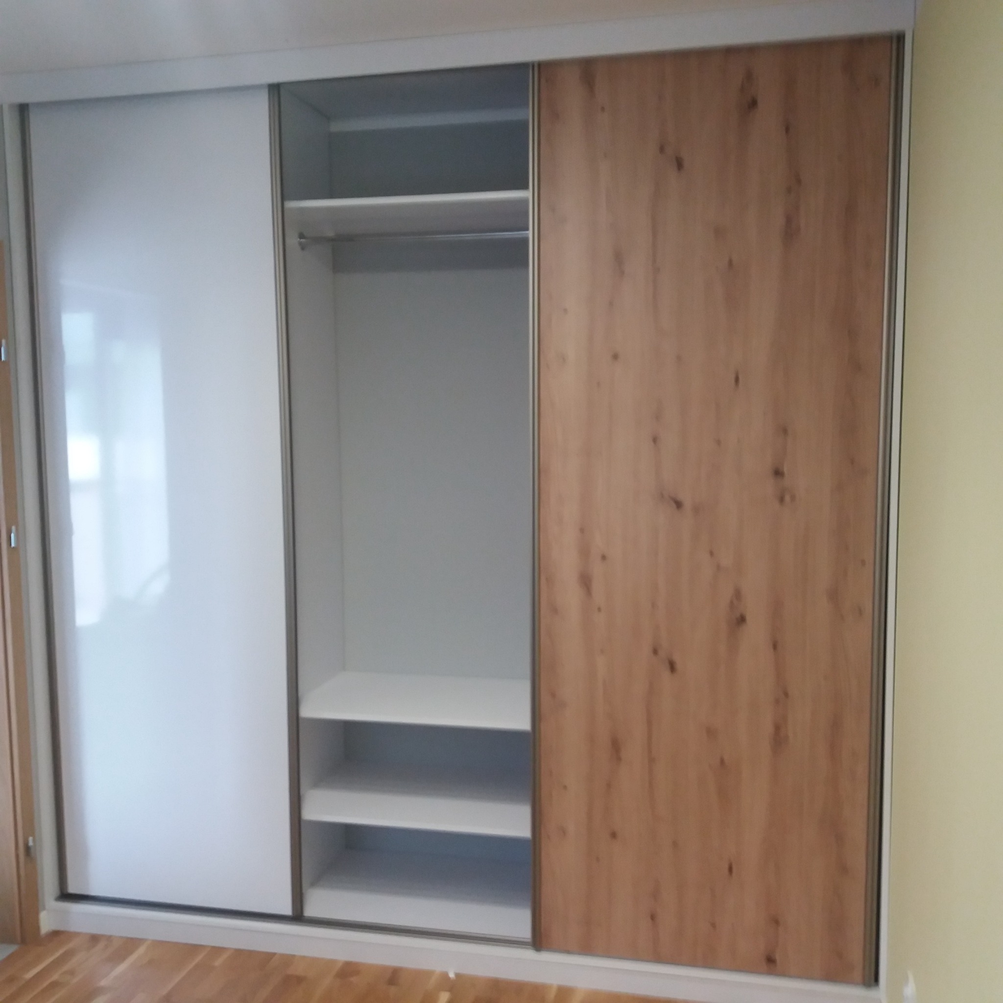 Wnętrze szafy wnękowej z przesuwnymi drzwiami: biały panel lustrzany i panel imitujący drewno, widoczne półki i drążek na ubrania.