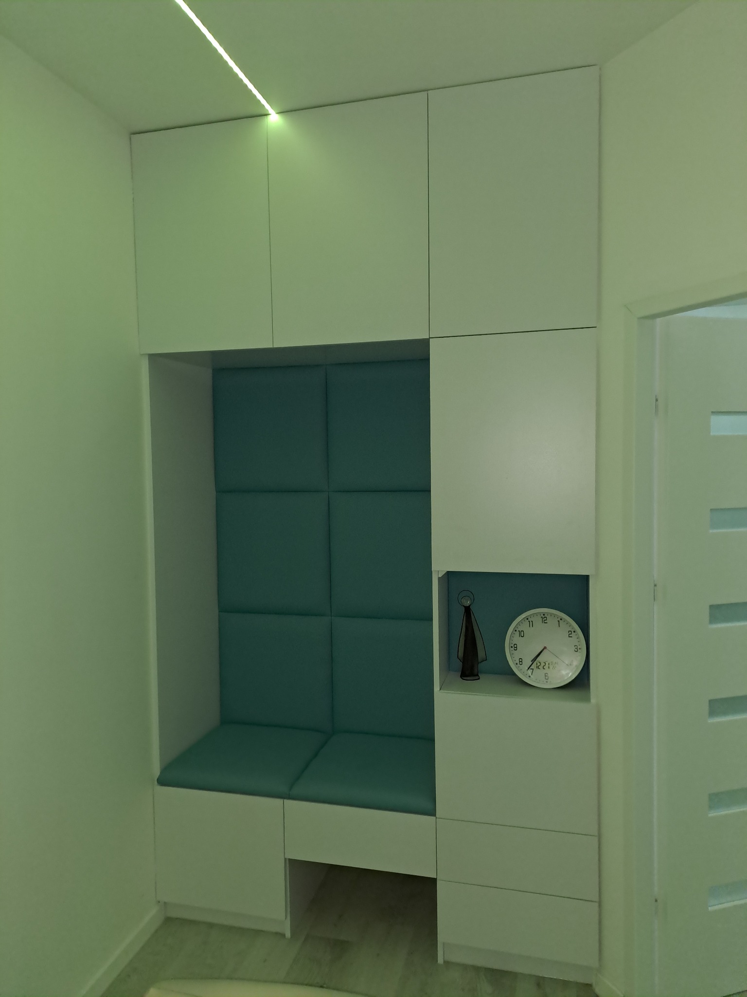 Biała zabudowa meblowa w przedpokoju z siedziskiem obitym turkusową tapicerką, oświetleniem LED i dekoracyjnym zegarem.