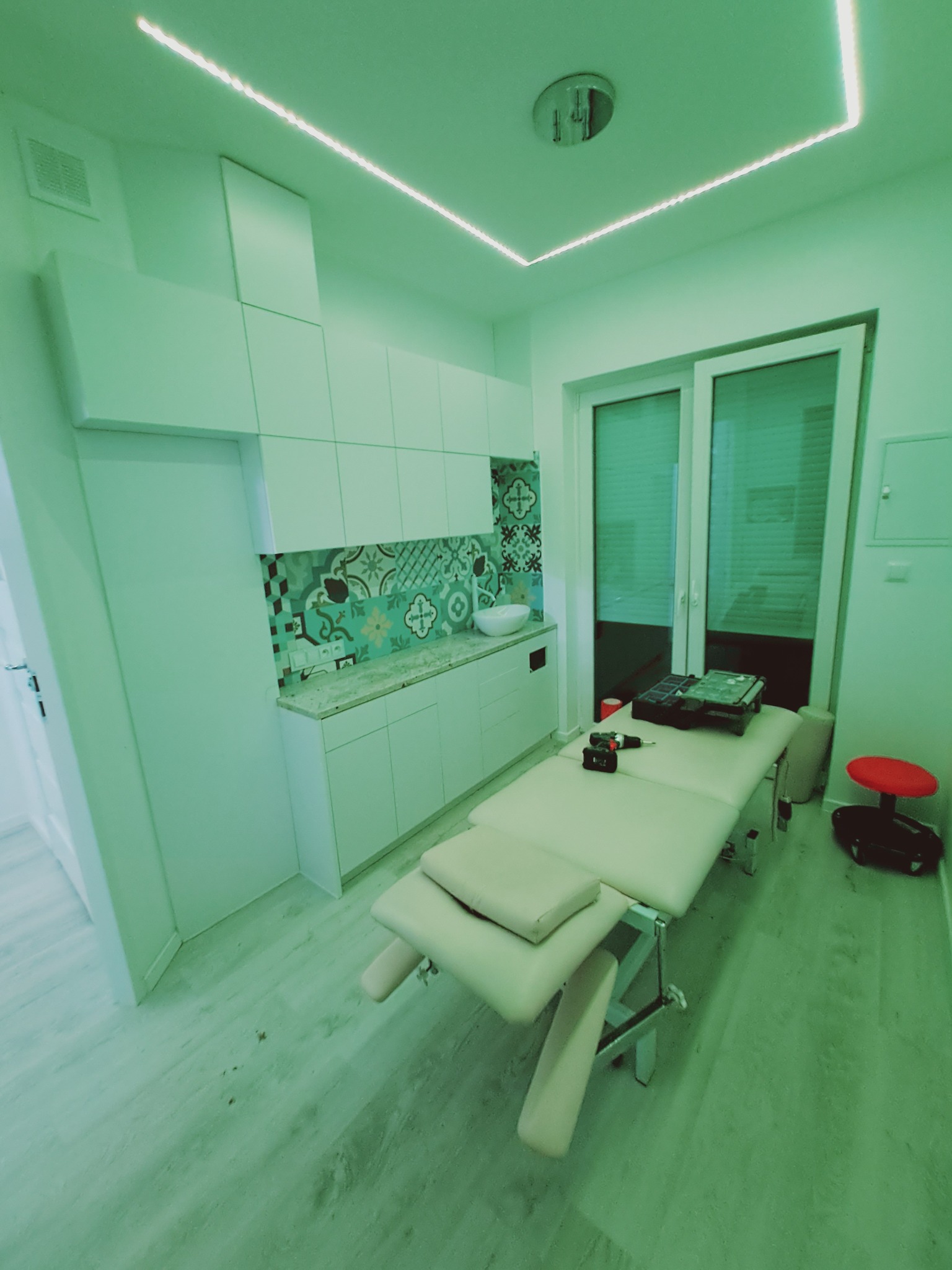 Wnętrze gabinetu z zabudową kuchenną, blatem z wzorzystymi płytkami i leżanką kosmetyczną, oświetlone ledowym paskiem na suficie.