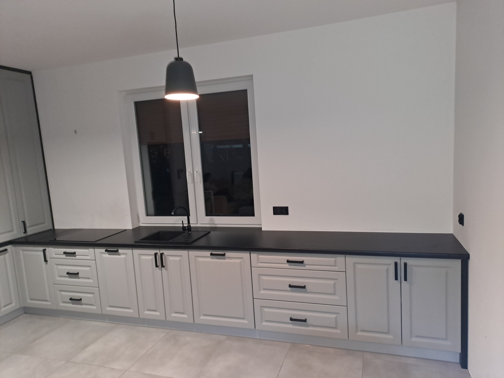 Jasnoszara zabudowa kuchenna z czarnym blatem i czarną armaturą, oświetlona wiszącą lampą w nowoczesnym stylu, widoczna z perspektywy podłogi.