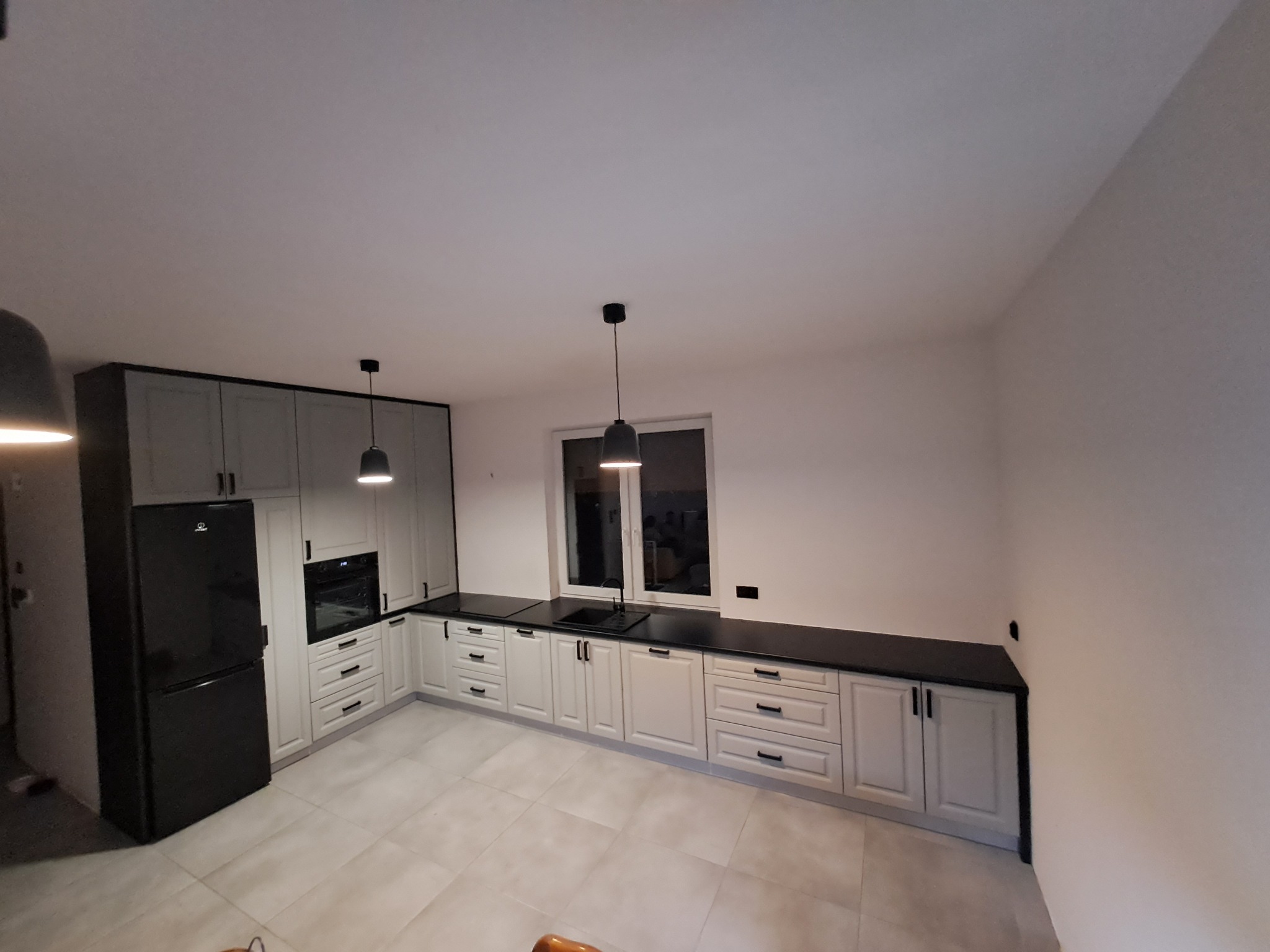 Jasna kuchnia z szarymi frontami i czarnymi blatami, oświetlona trzema lampami wiszącymi, z czarną lodówką i oknem w tle.