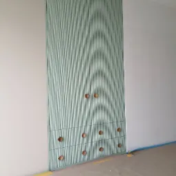 Wbudowana szafa z ryflowanymi frontami w pastelowym odcieniu zieleni, z drewnianymi okrągłymi uchwytami, umieszczona między białymi ścianami, podłoga zabezpieczona taśmą malarską.