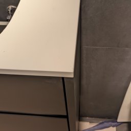 Fabryka Mebli Farmahei - Fragment łazienki z umywalką nablatową na szafce z szufladami, widoczne szare płytki na ścianie i silikon zabezpieczający połączenie szafki ze ścianą.