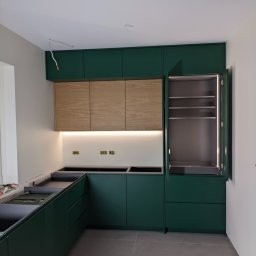 Fabryka Mebli Farmahei - Wnętrze kuchni w trakcie montażu, z zielonymi szafkami dolnymi i górnymi z drewnianymi frontami, podświetleniem LED pod szafkami górnymi oraz otwartą szafką z oświetleniem wewnętrznym. Widoczne...