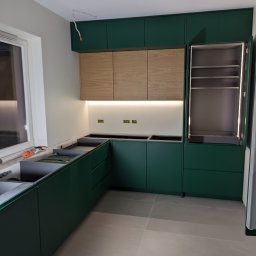 Fabryka Mebli Farmahei - Wnętrze kuchni w trakcie montażu: zielone szafki dolne i górne, drewniane szafki wiszące podświetlone listwą LED, otwarty słupek z oświetleniem wewnętrznym, widoczne instalacje elektryczne, szare...
