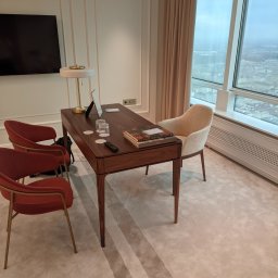 Fabryka Mebli Farmahei - Nowoczesne biuro w apartamencie z drewnianym biurkiem, krzesłami w odcieniach czerwieni i beżu, lampą, książkami i widokiem na miasto przez duże okno.