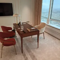 Nowoczesne biuro w apartamencie z drewnianym biurkiem, krzesłami w odcieniach czerwieni i beżu, lampą, książkami i widokiem na miasto przez duże okno.