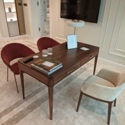 Fabryka Mebli Farmahei - Nowoczesne biurko z ciemnego drewna z dwiema szufladami, otoczone trzema krzesłami, z książkami, pilotem i szklankami na blacie, a także lampką i ramką na zdjęcia, na tle telewizora i otwartych drzwi.