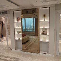 Fabryka Mebli Farmahei - Nowoczesna garderoba z lustrzanymi drzwiami przesuwnymi, oświetlonymi półkami z dekoracjami i widokiem na miasto w tle.