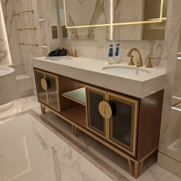 Fabryka Mebli Farmahei - Nowoczesna szafka łazienkowa z dwoma umywalkami, złotymi bateriami i okrągłymi uchwytami na tle marmurowych ścian i podłóg, podświetlane lustro.